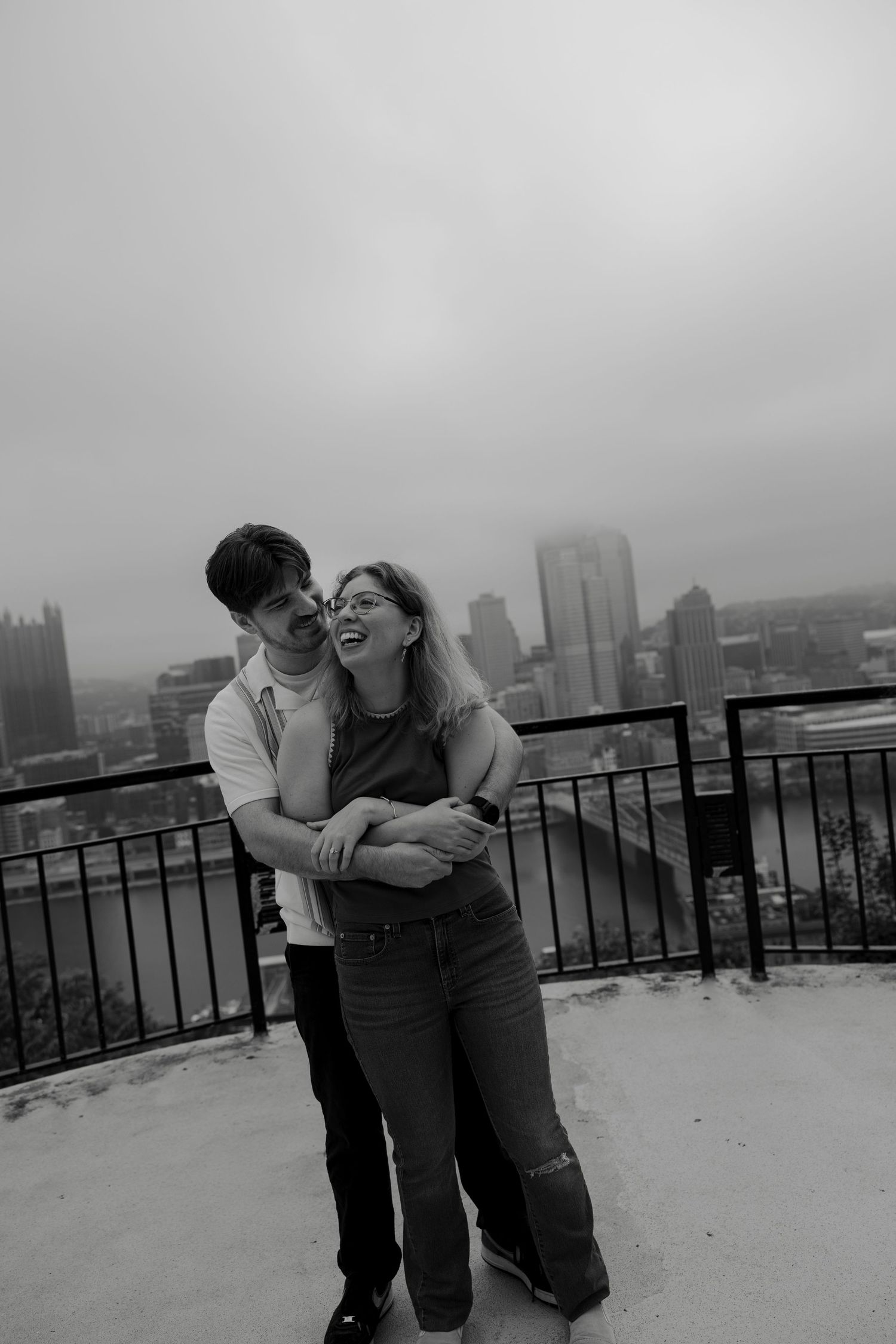 Black and white photo of a joyful couple embracing on a snowy overlook with a city skyline behind them.