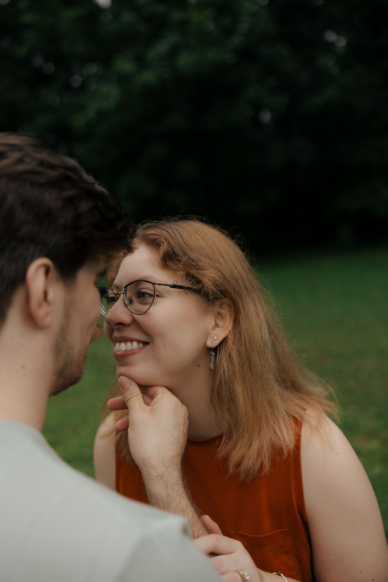 Smiling woman with glasses and auburn hair gazes lovingly at her partner during an outdoor couples session.