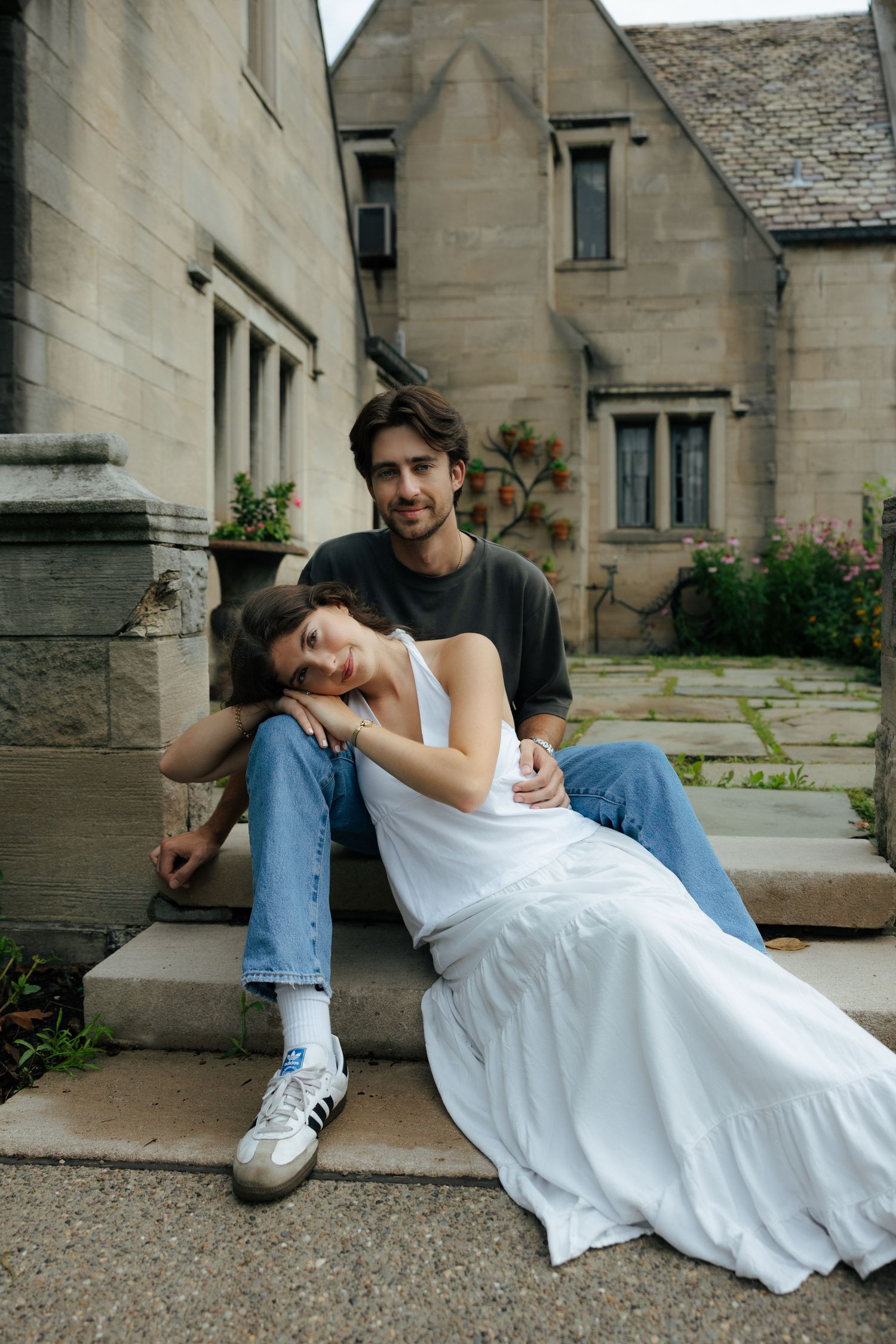 Couple sitting on stone steps, woman in white dress leaning on man's knee outdoors.