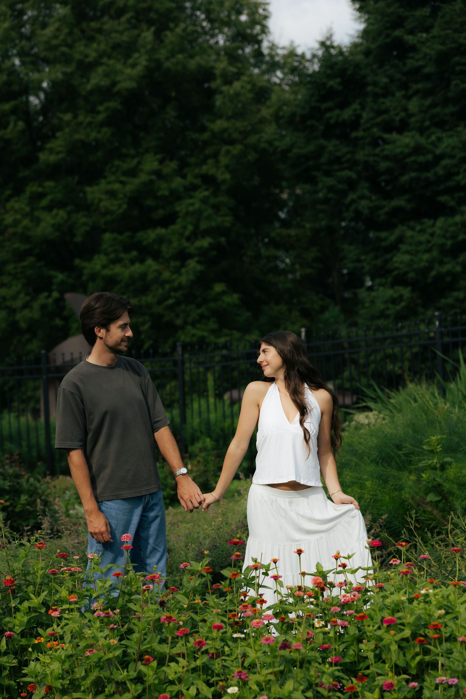 A couple holds hands while walking through a vibrant flower garden with lush green trees.
