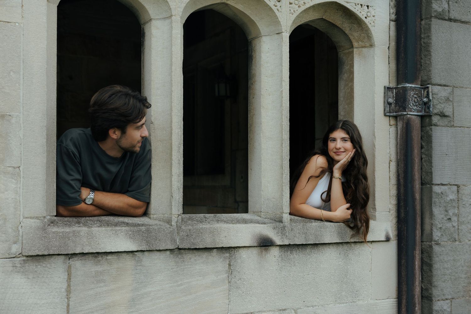 A smiling couple leaning out of separate stone arch windows of a historic building.