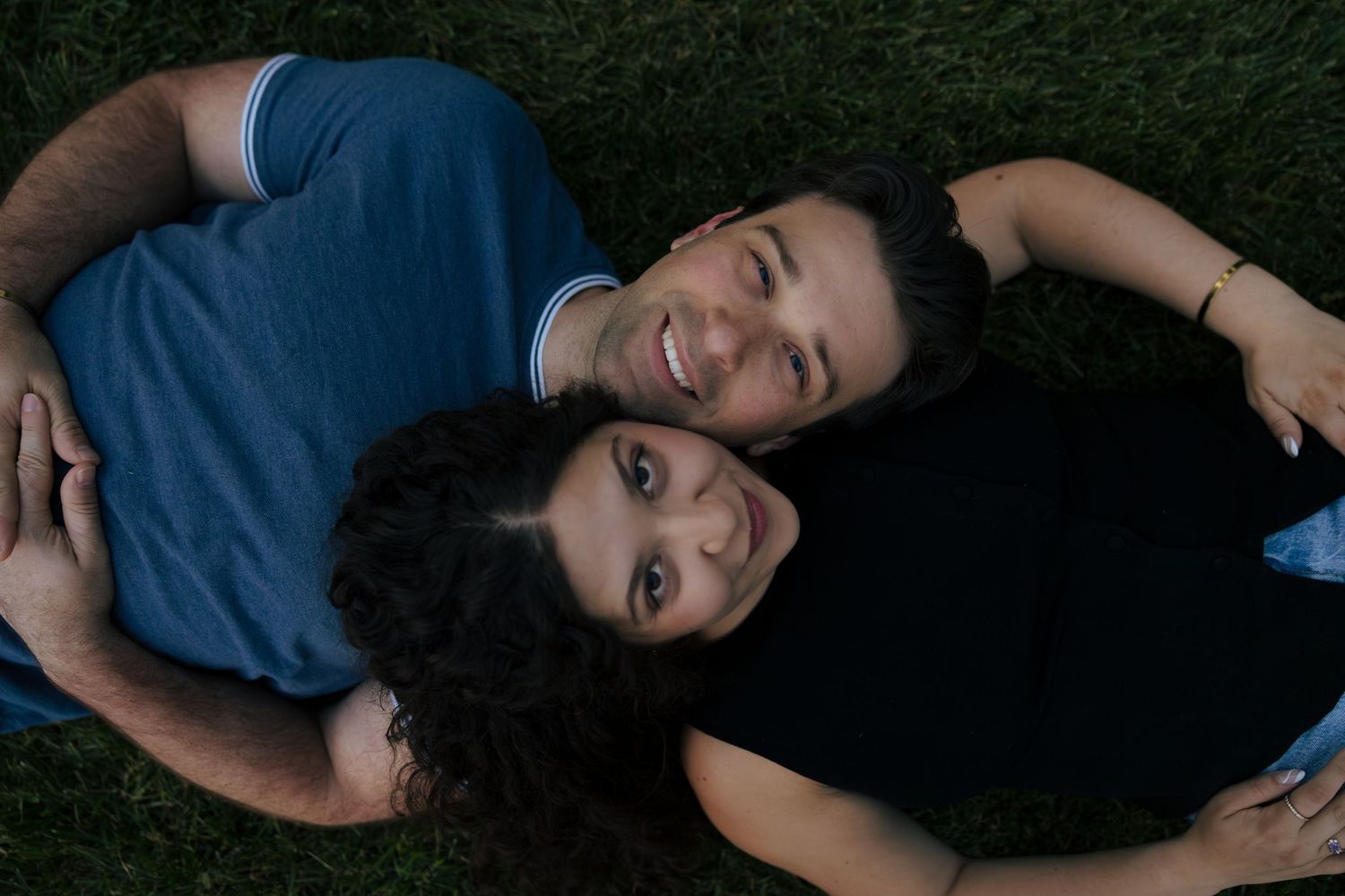 A smiling couple lying on green grass, looking up at the camera in a romantic outdoor portrait.