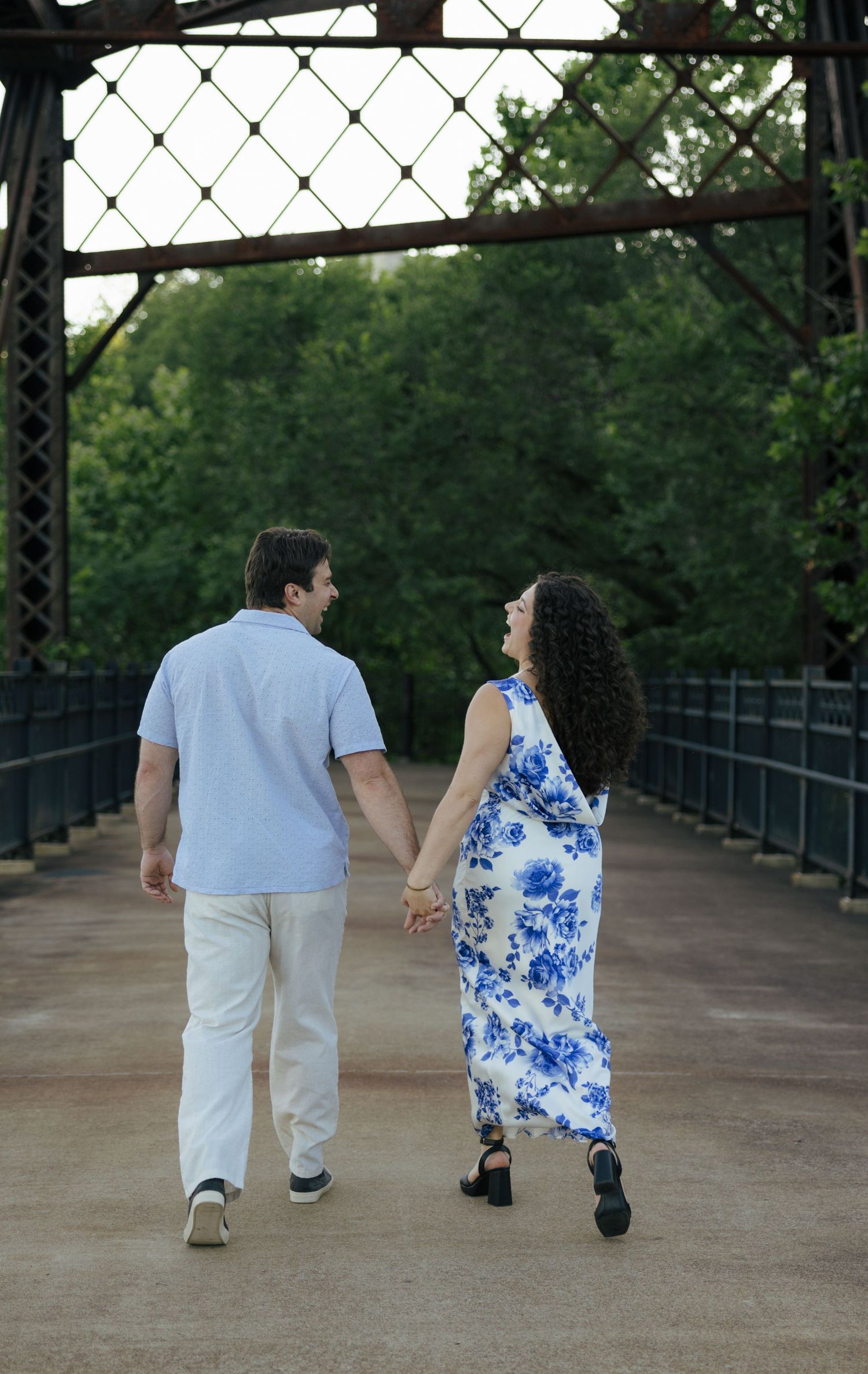 A couple holds hands and walks together on a rustic iron bridge, smiling at each other.