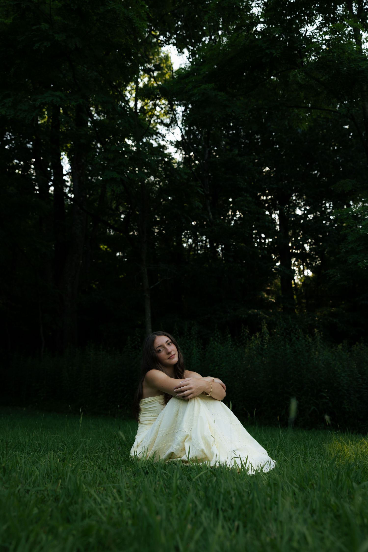 Young woman in white gown sitting on green grass in a dark forest setting, hugging her knees contemplatively.