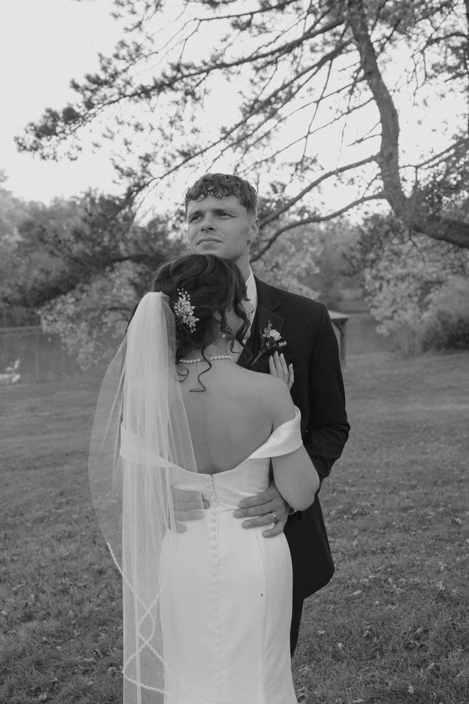 Groom embracing bride from behind in black and white outdoor wedding portrait among trees.