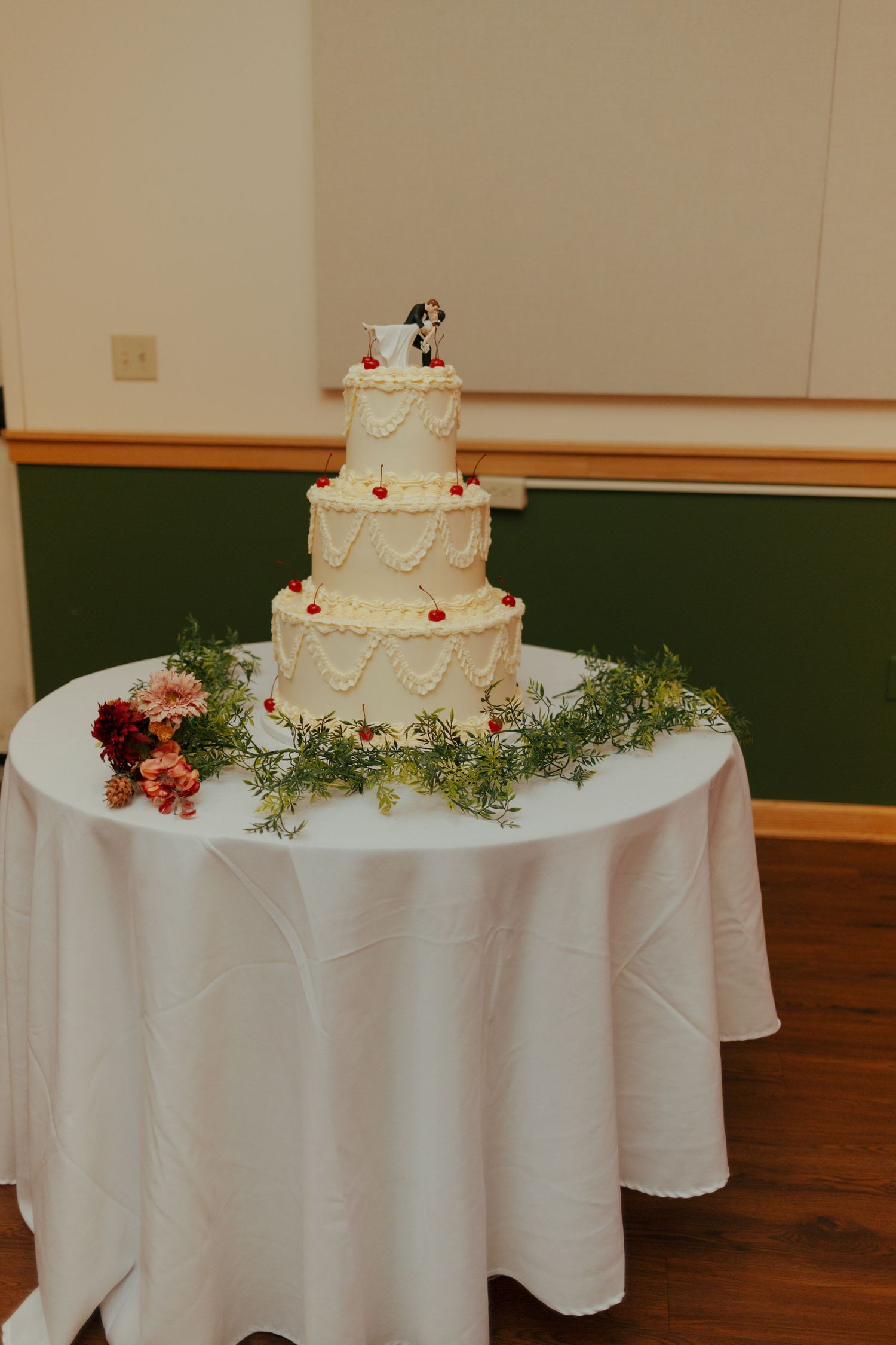 Four-tier white wedding cake with pearl details and couple topper on draped table.