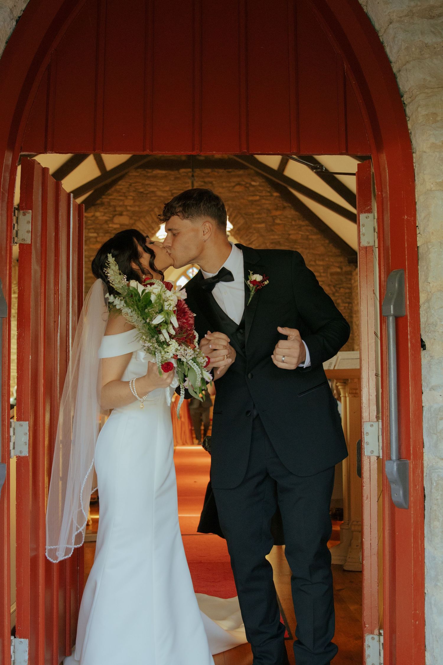 Bride and groom share a kiss at the entrance of a rustic church with red arched doors on their wedding day.