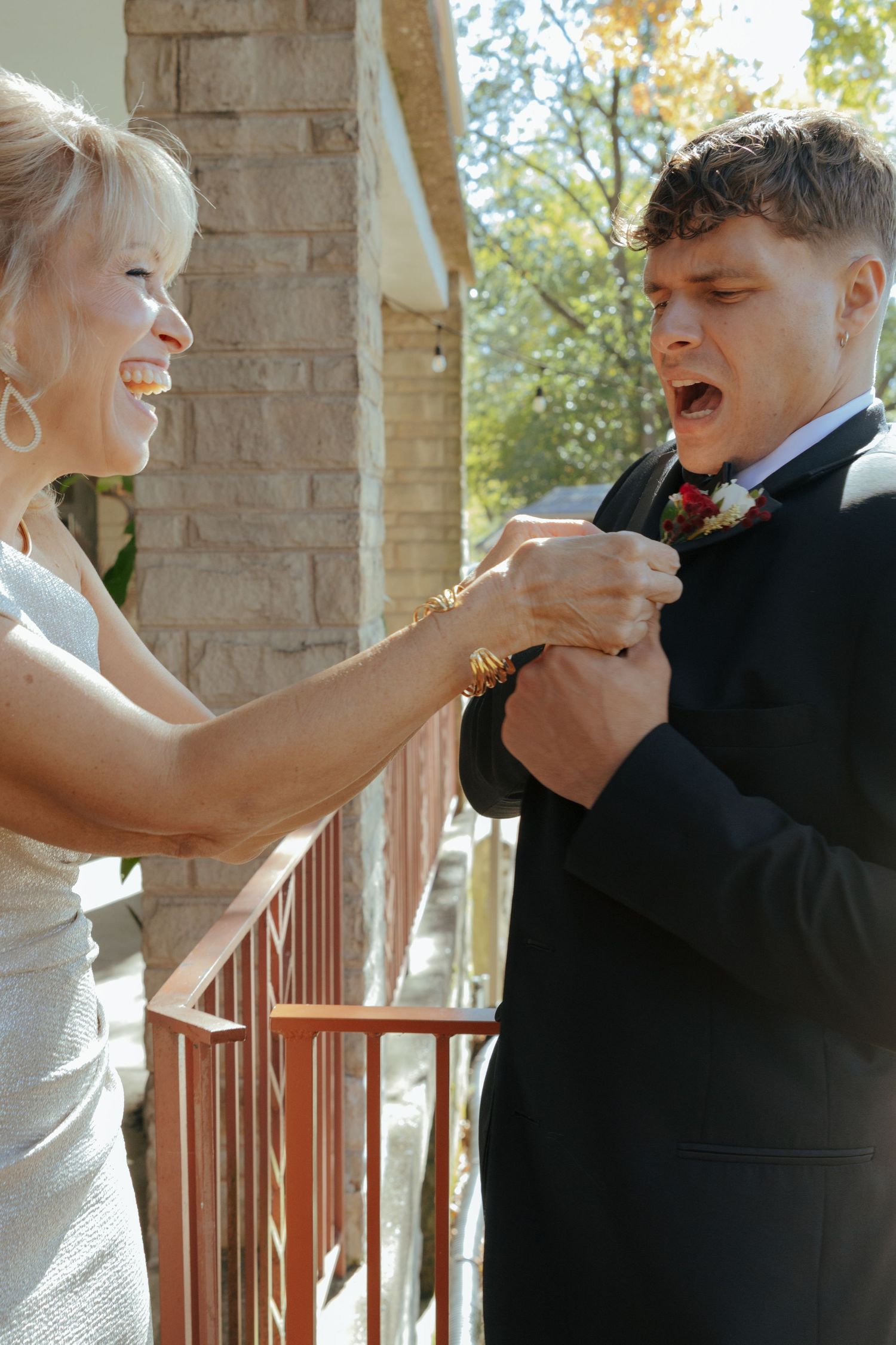 Woman smiling while adjusting boutonniere on groom in black suit on outdoor porch before wedding.