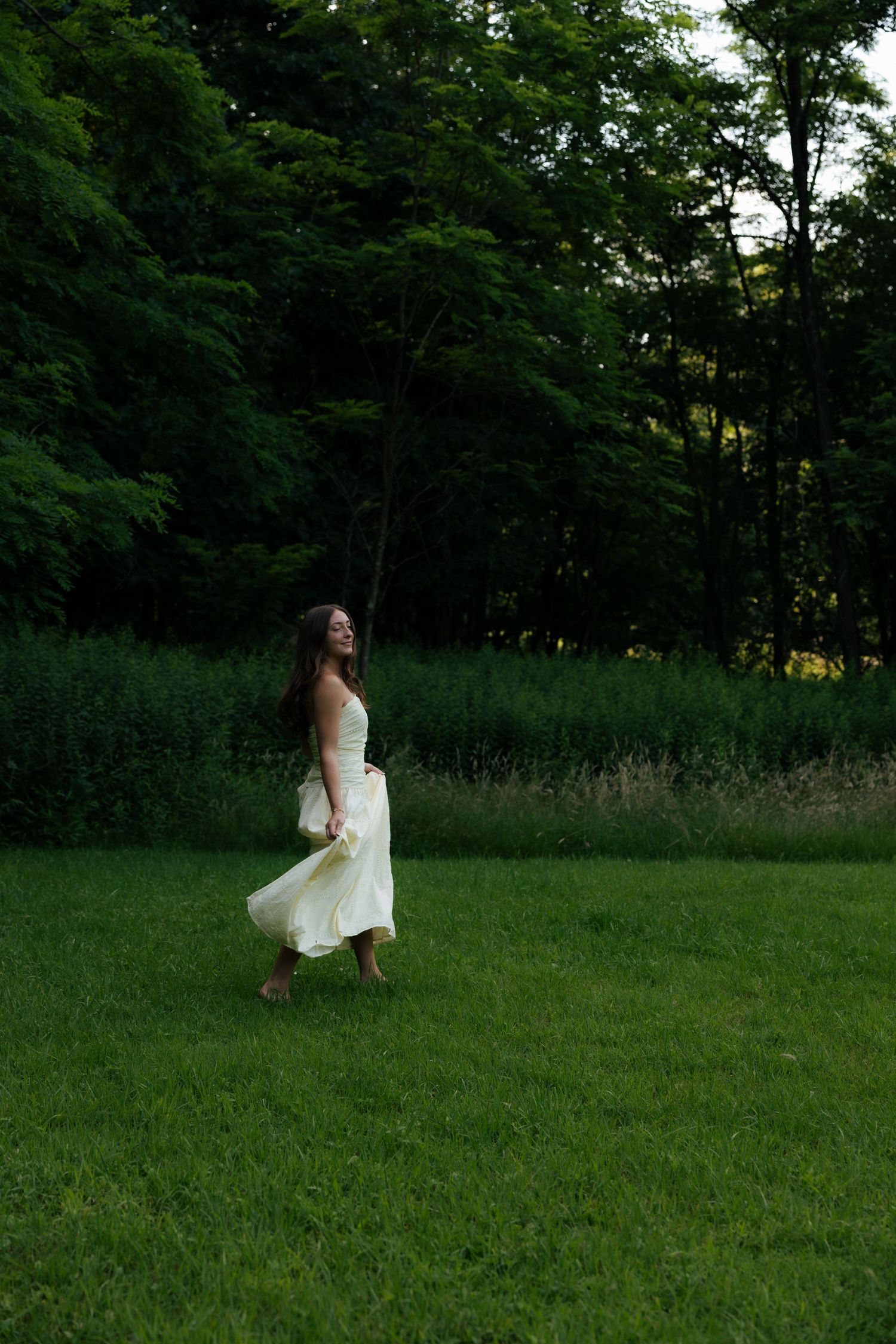 A woman in a flowing white dress twirls barefoot on lush green grass with trees in the background.