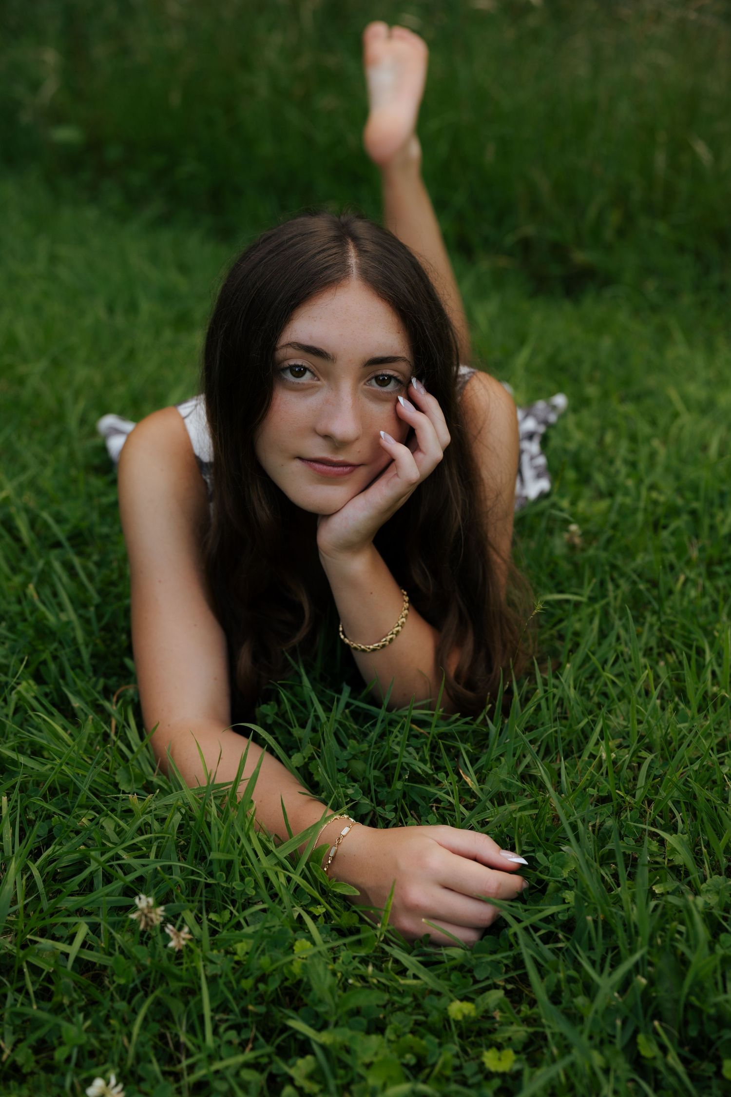 Young woman with long dark hair lying in lush green grass, resting chin on hand, wearing a bracelet and anklet.