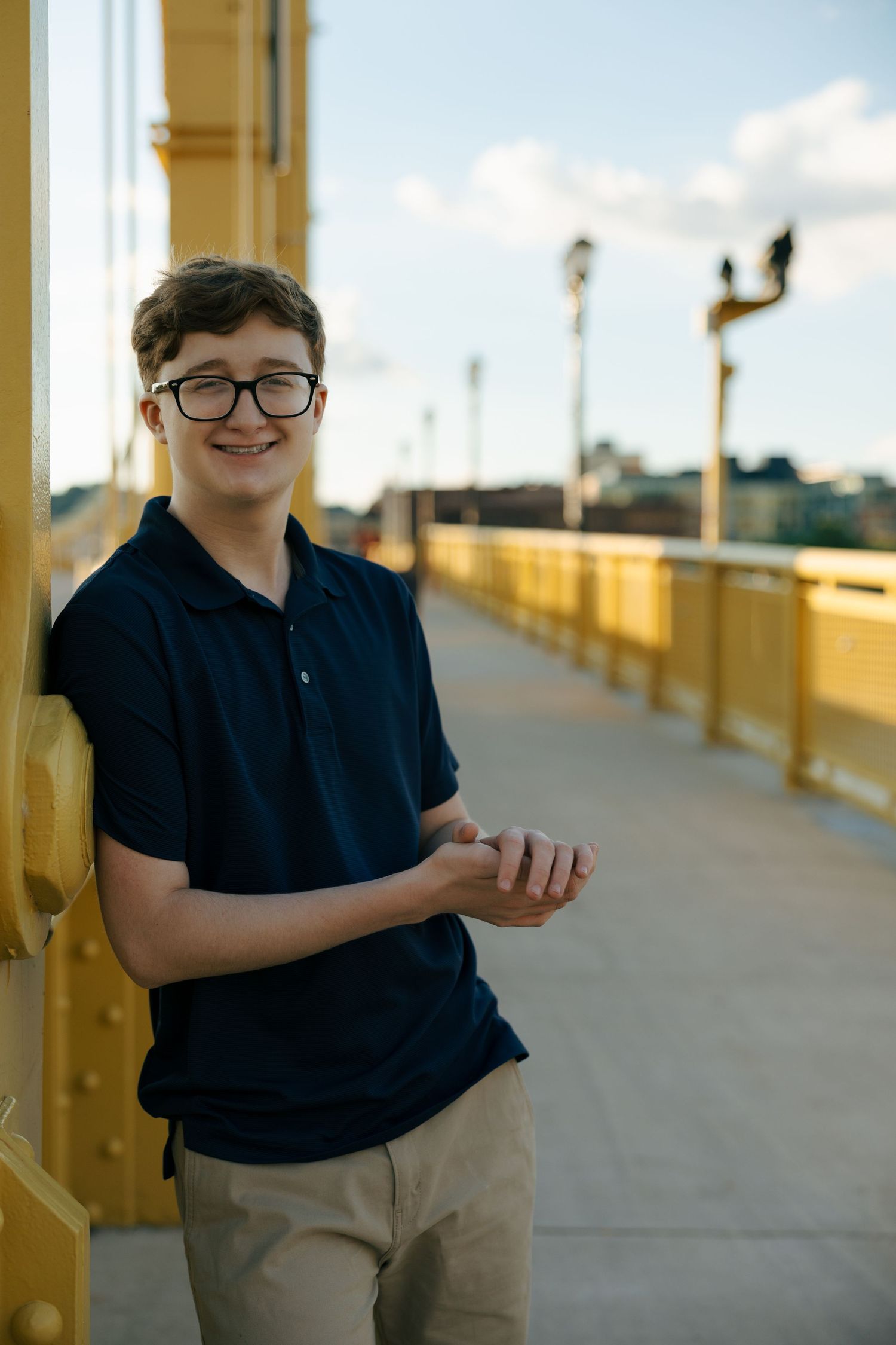 Smiling teen in navy polo leaning on yellow bridge pillar.