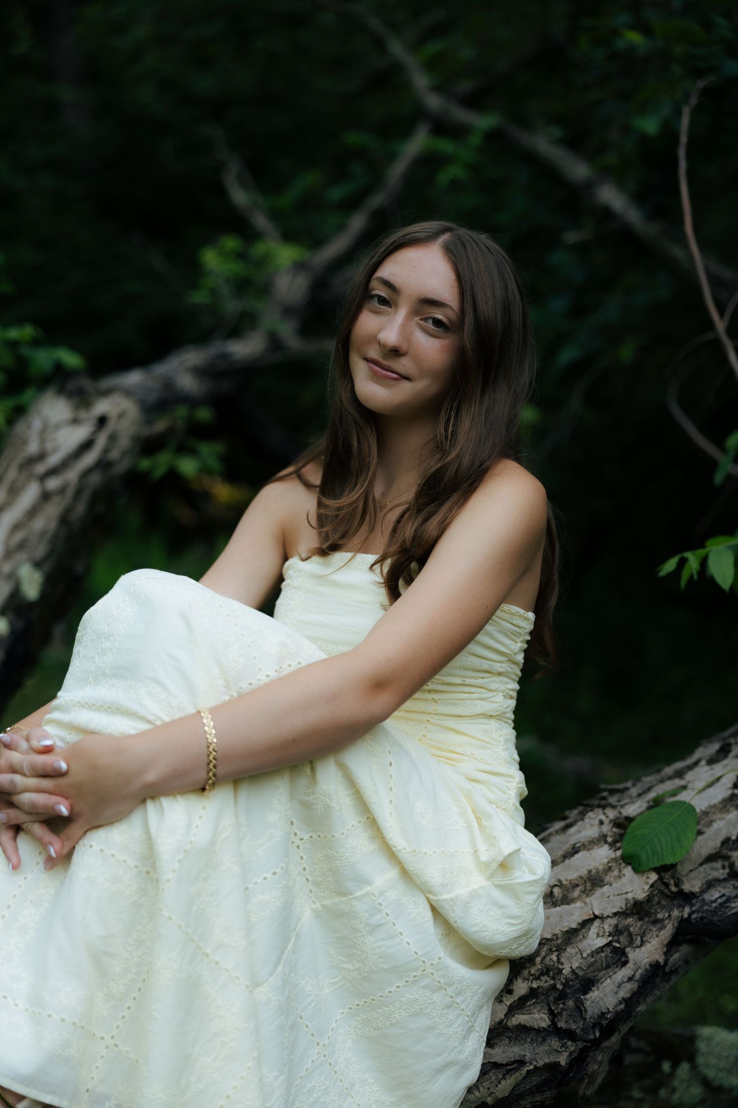 Young woman in a cream strapless dress sitting gracefully on a log in a lush green forest setting.