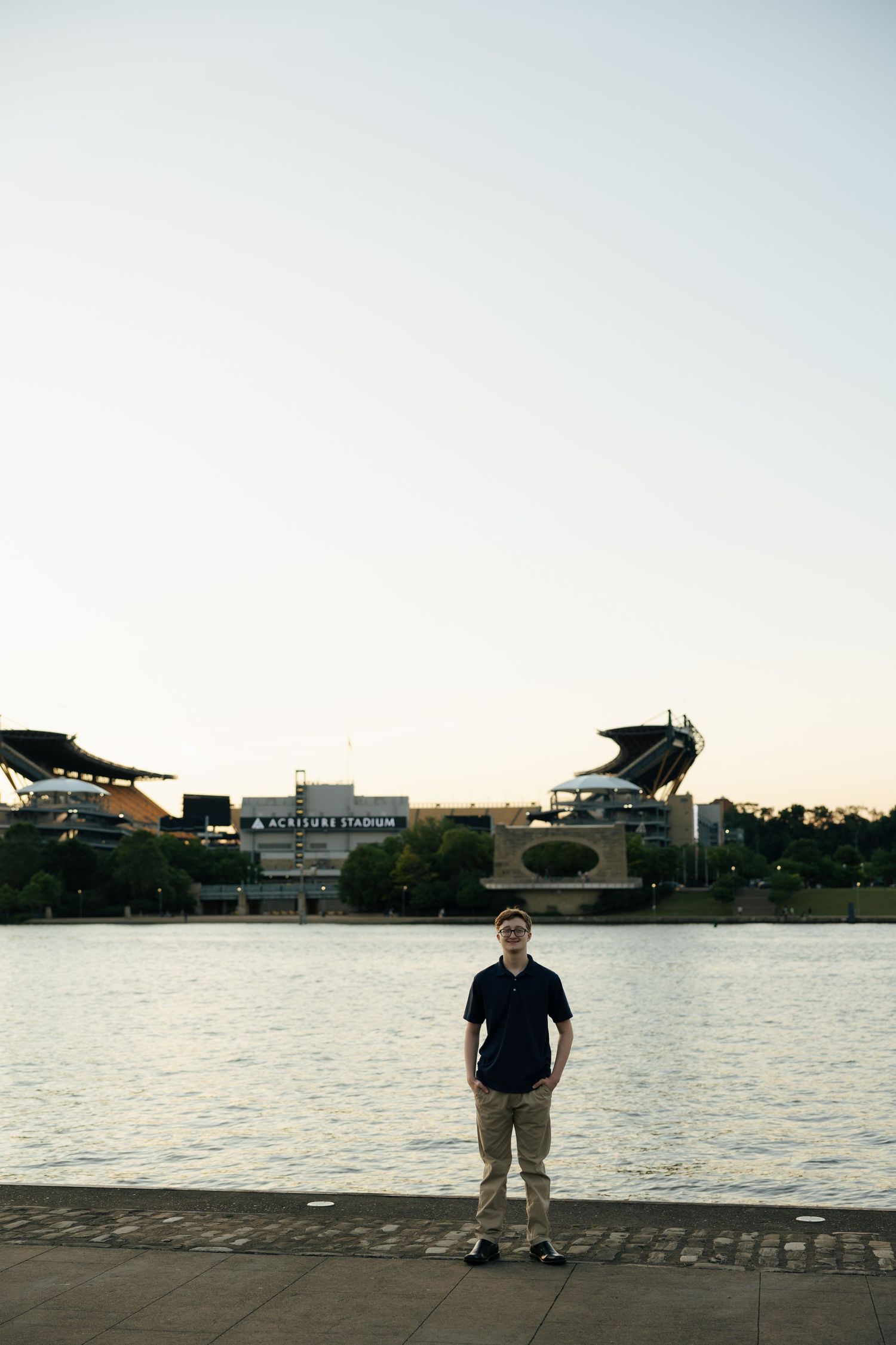 Teen boy in navy polo standing on Pittsburgh riverfront with Acrisure Stadium across the water.