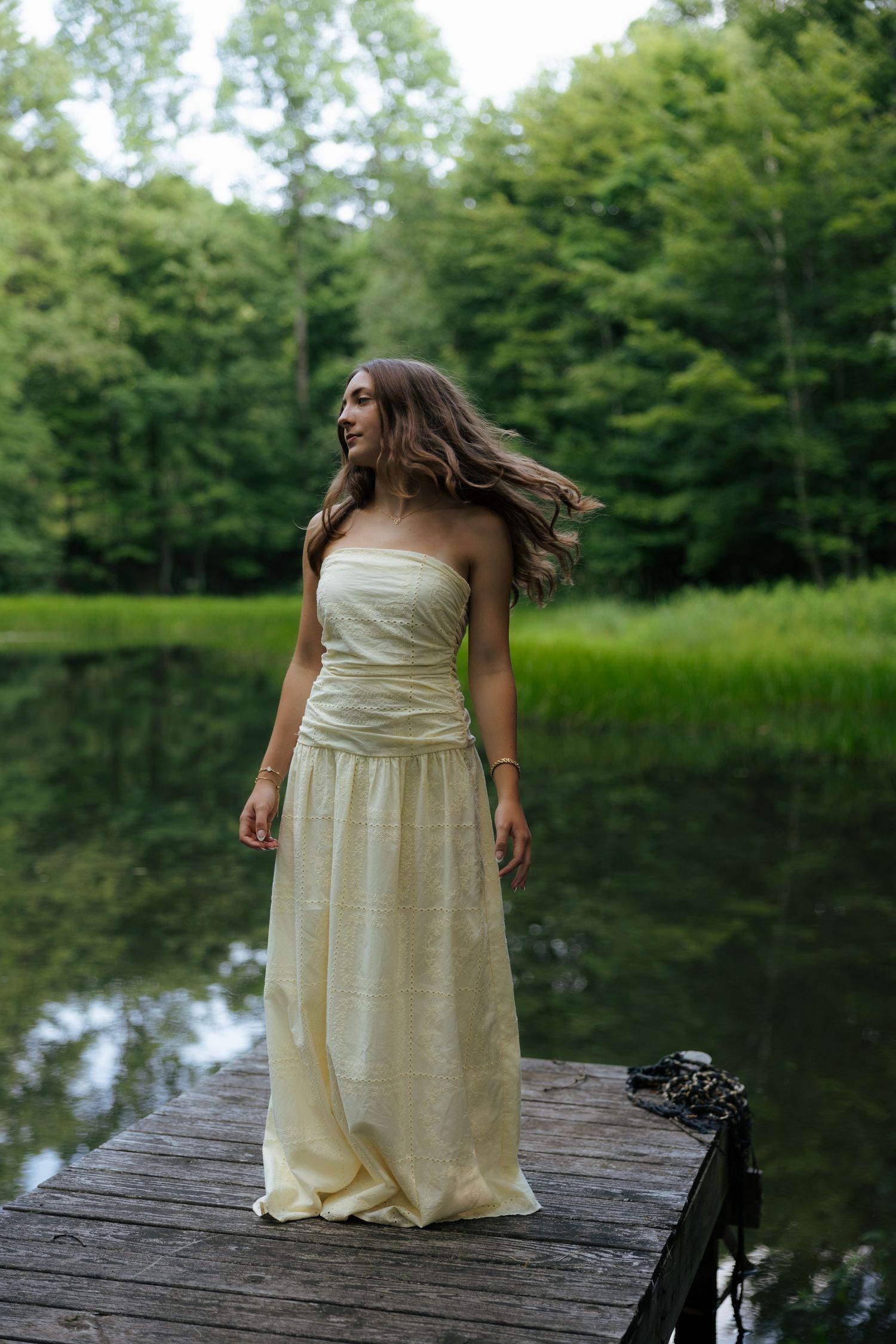 A young woman in a cream strapless gown stands on a wooden dock by a calm forest lake.