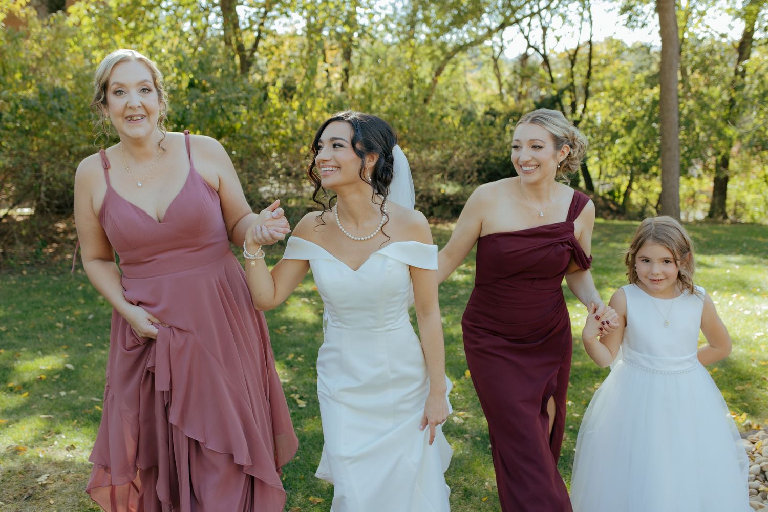Bride in white gown walks with bridesmaids in dusty rose and burgundy dresses and flower girl in white outdoors.