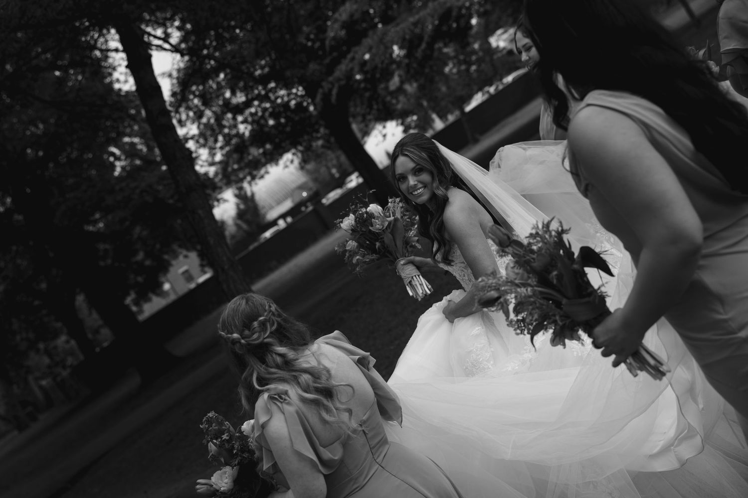 Black and white wedding photo of a smiling bride in a white gown holding a bouquet, surrounded by flower girls outdoors.