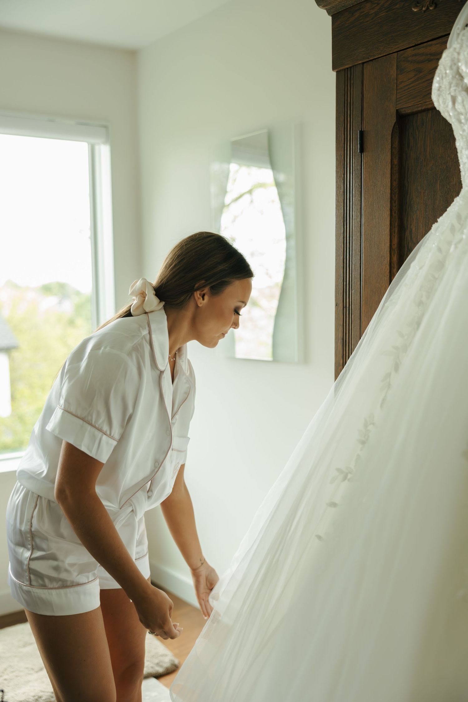 Bride in white shorts examining her lace wedding gown hanging on a wooden wardrobe in a bright room.