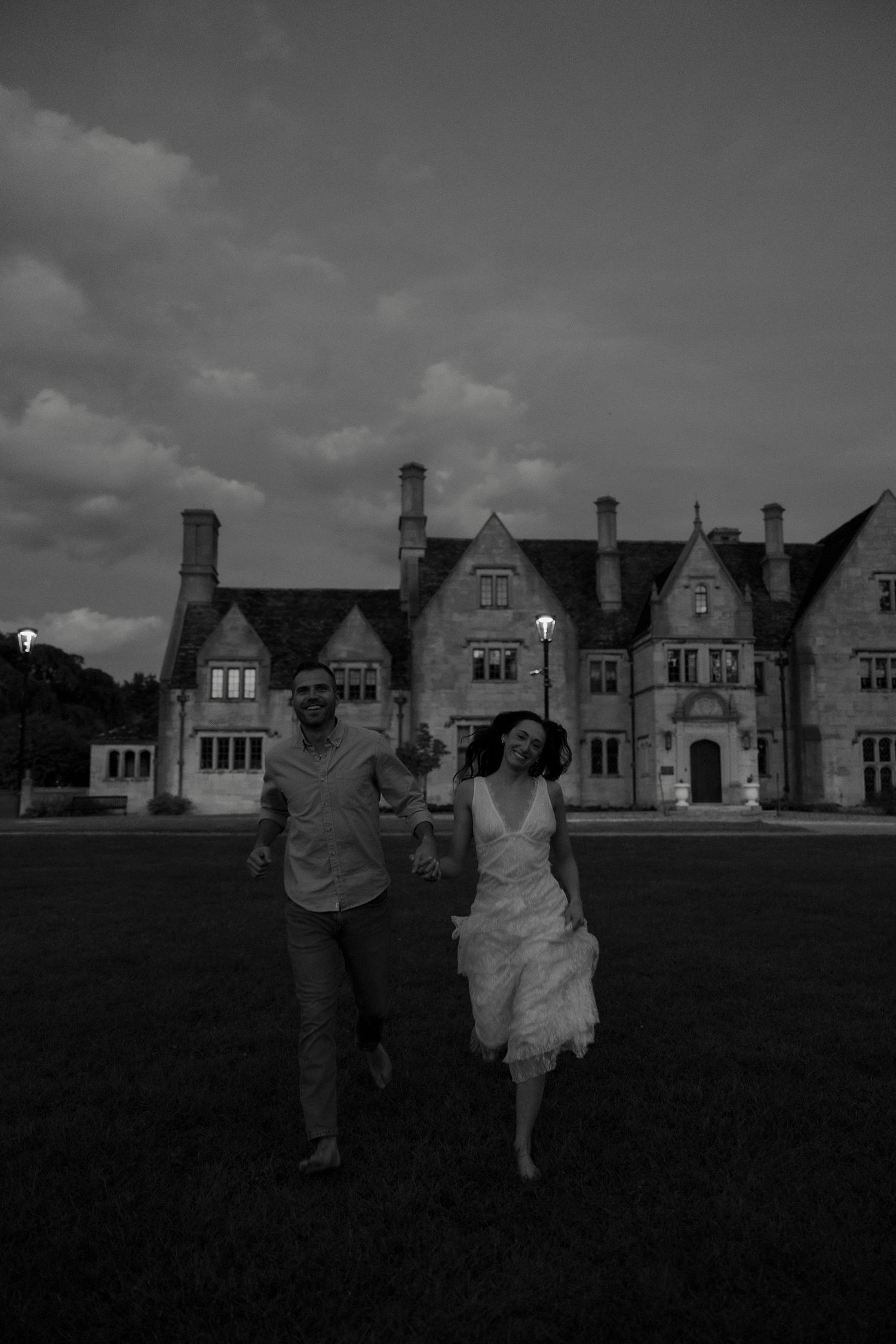 Black and white photo of couple running hand in hand before grand stone manor at dusk.