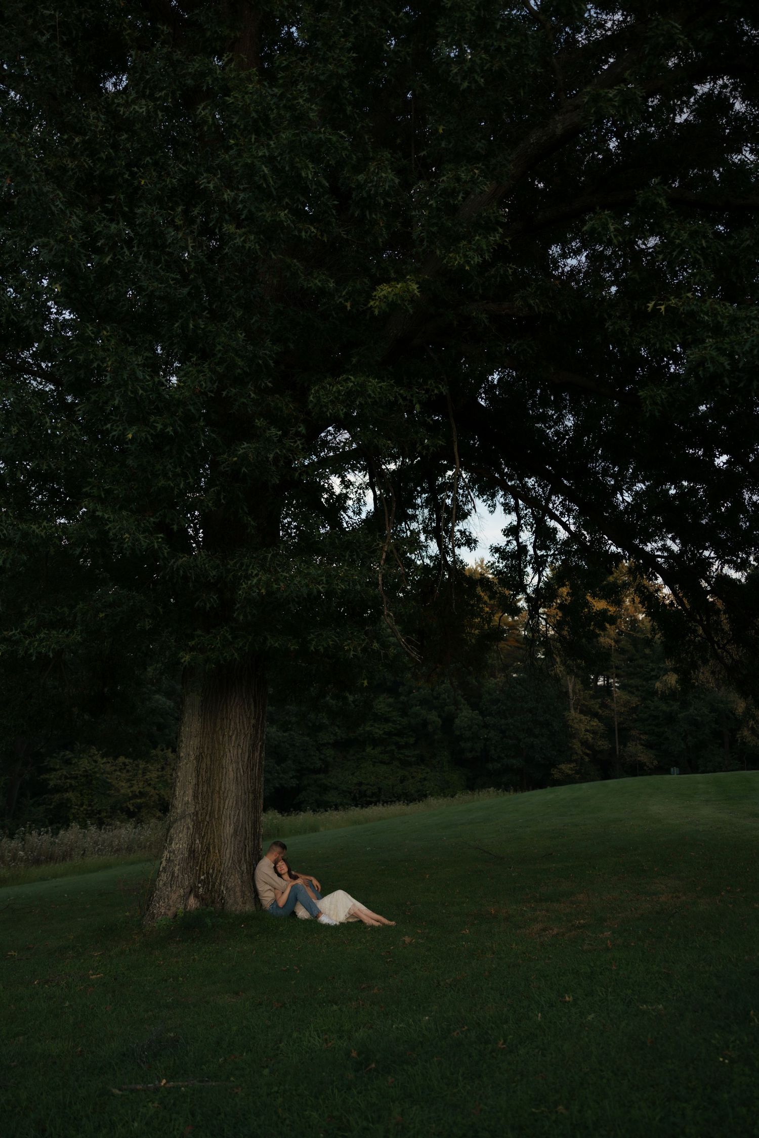 Couple sitting together leaning against a large tree trunk in a dark, lush green park at dusk.