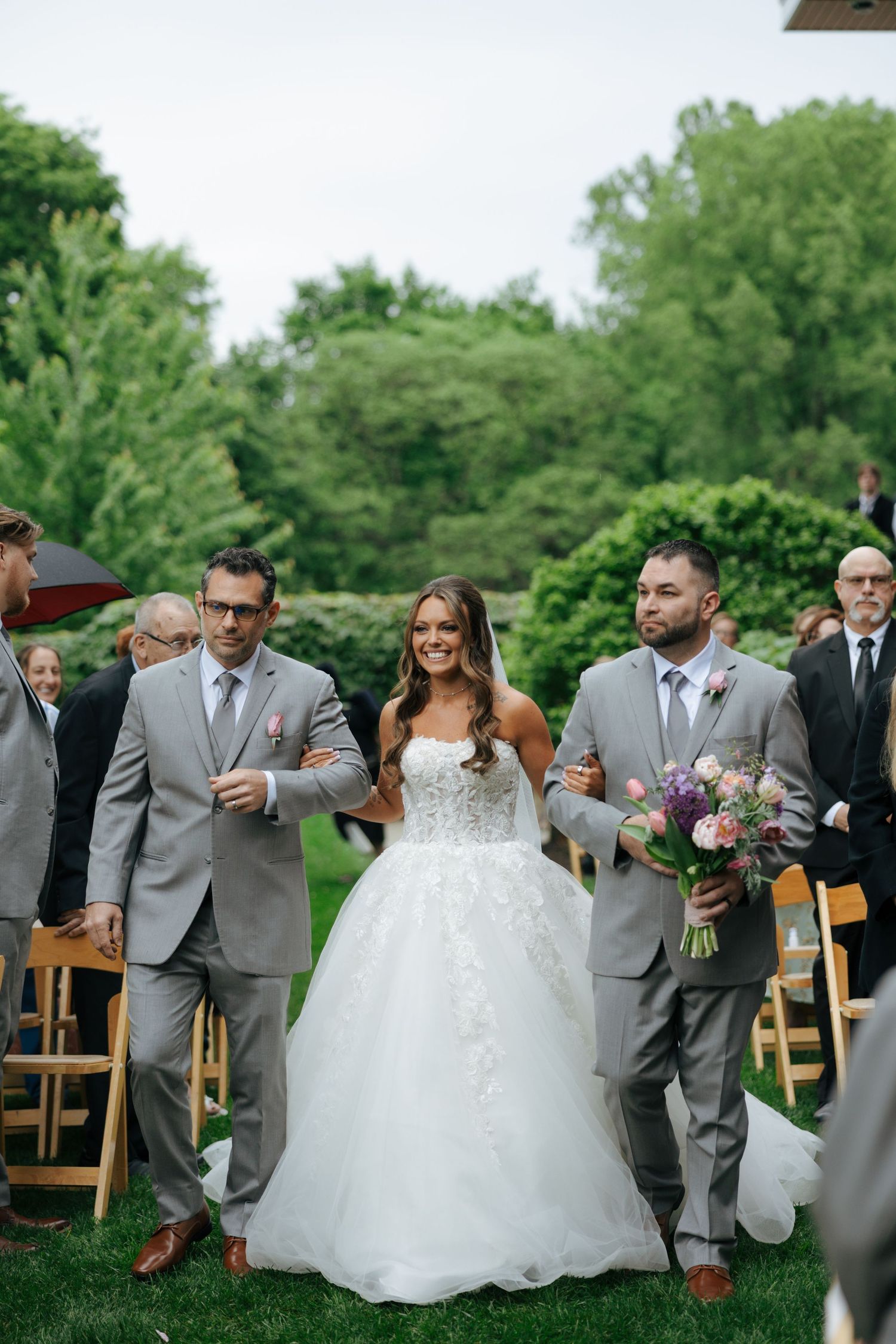 Bride walks down outdoor wedding aisle in white lace ballgown, escorted by two men in gray suits.