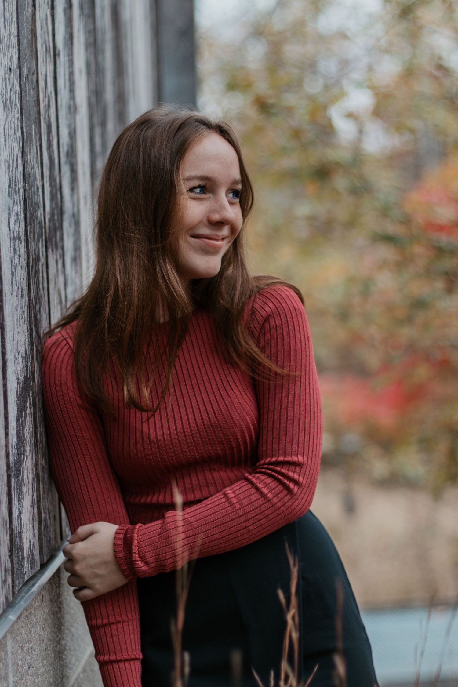 Person in red sweater poses against wooden post in autumn setting.