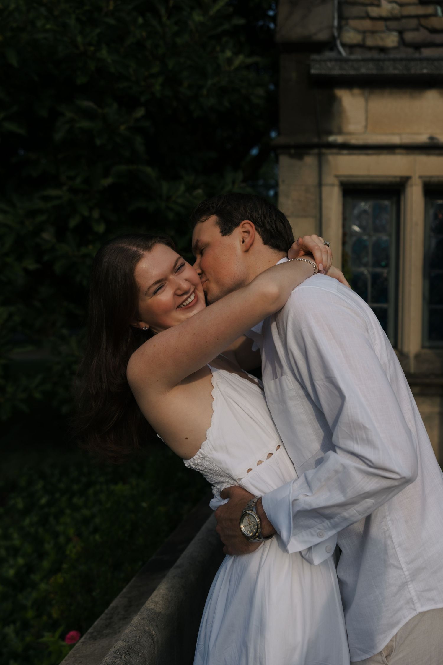 Laughing woman in white dress hugging man in white shirt outdoors near historic stone building.