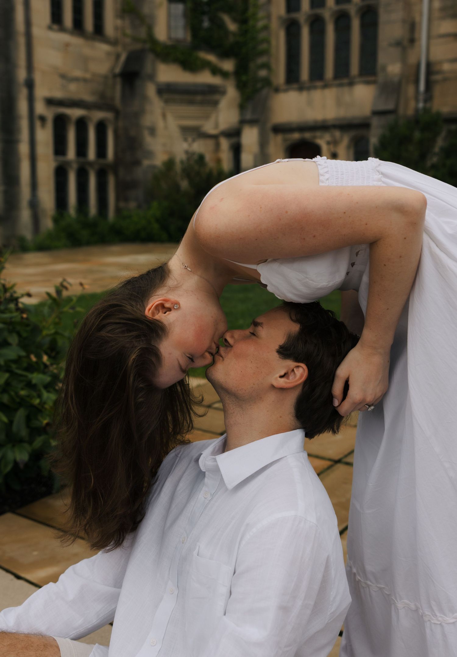 Couple sharing a romantic upside-down kiss outside a historic stone building.