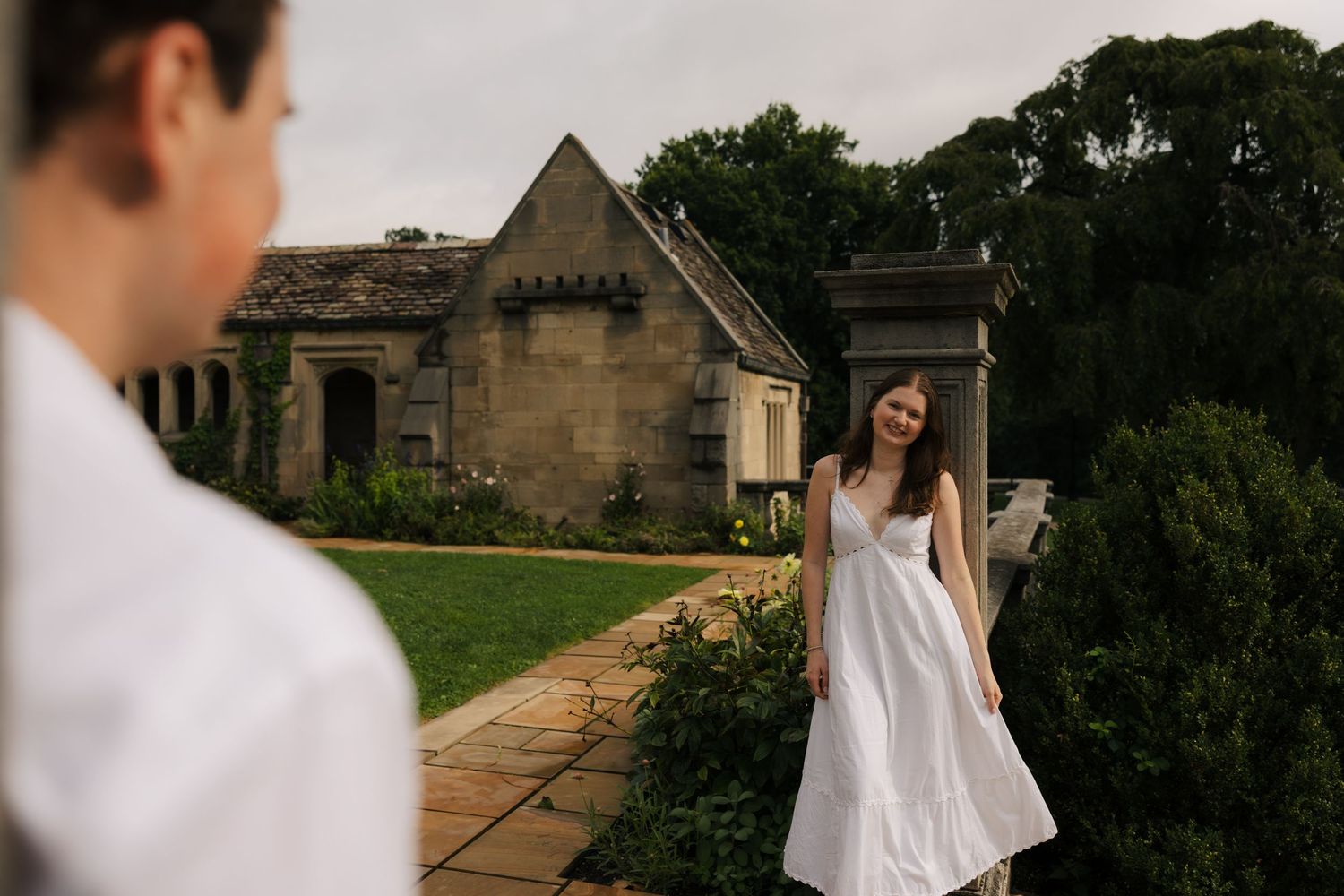 Woman in white dress smiling at man outdoors near a stone cottage garden.