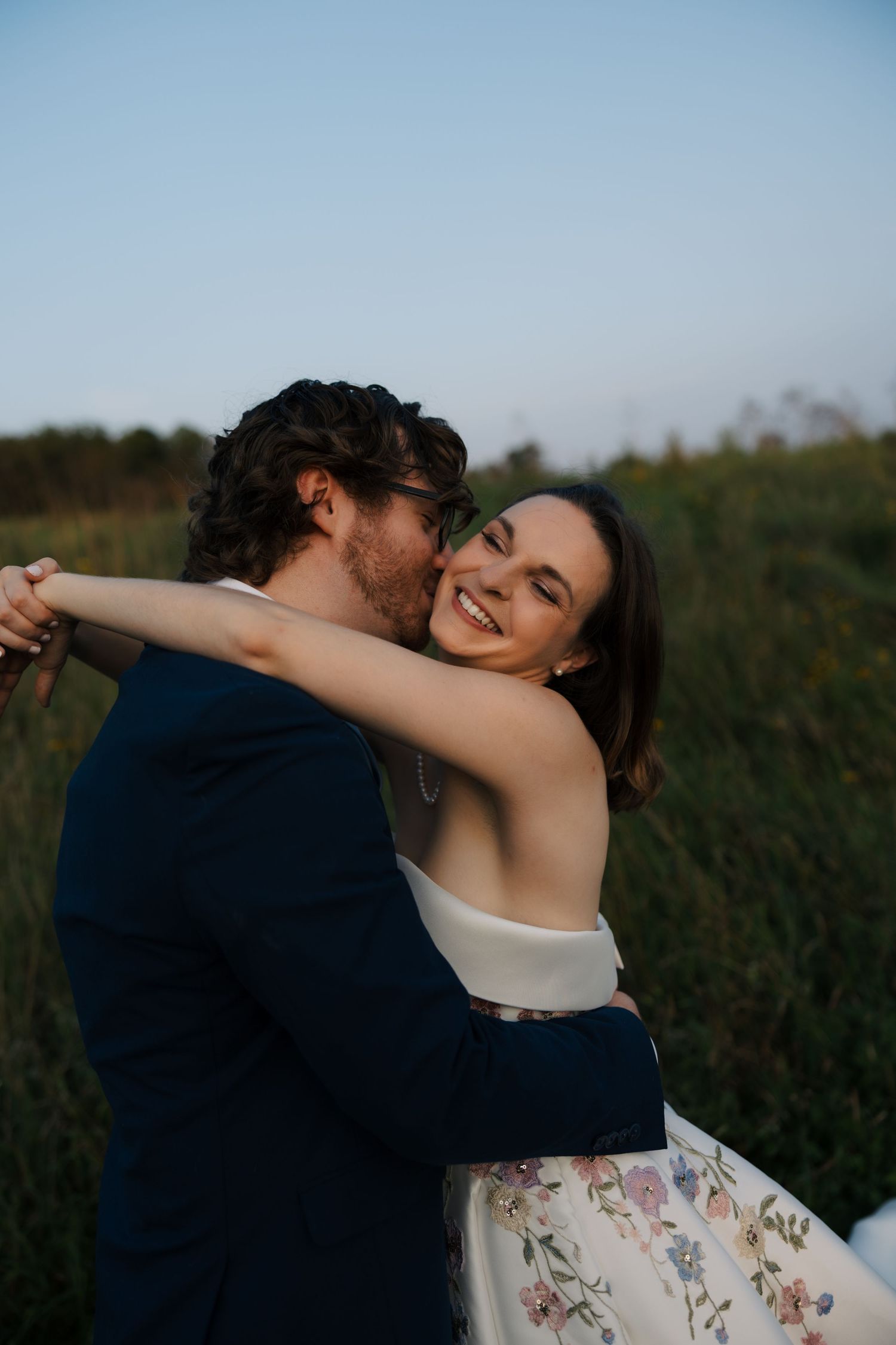 Groom in navy suit embraces laughing bride in floral wedding gown in a sunlit green meadow at sunset.
