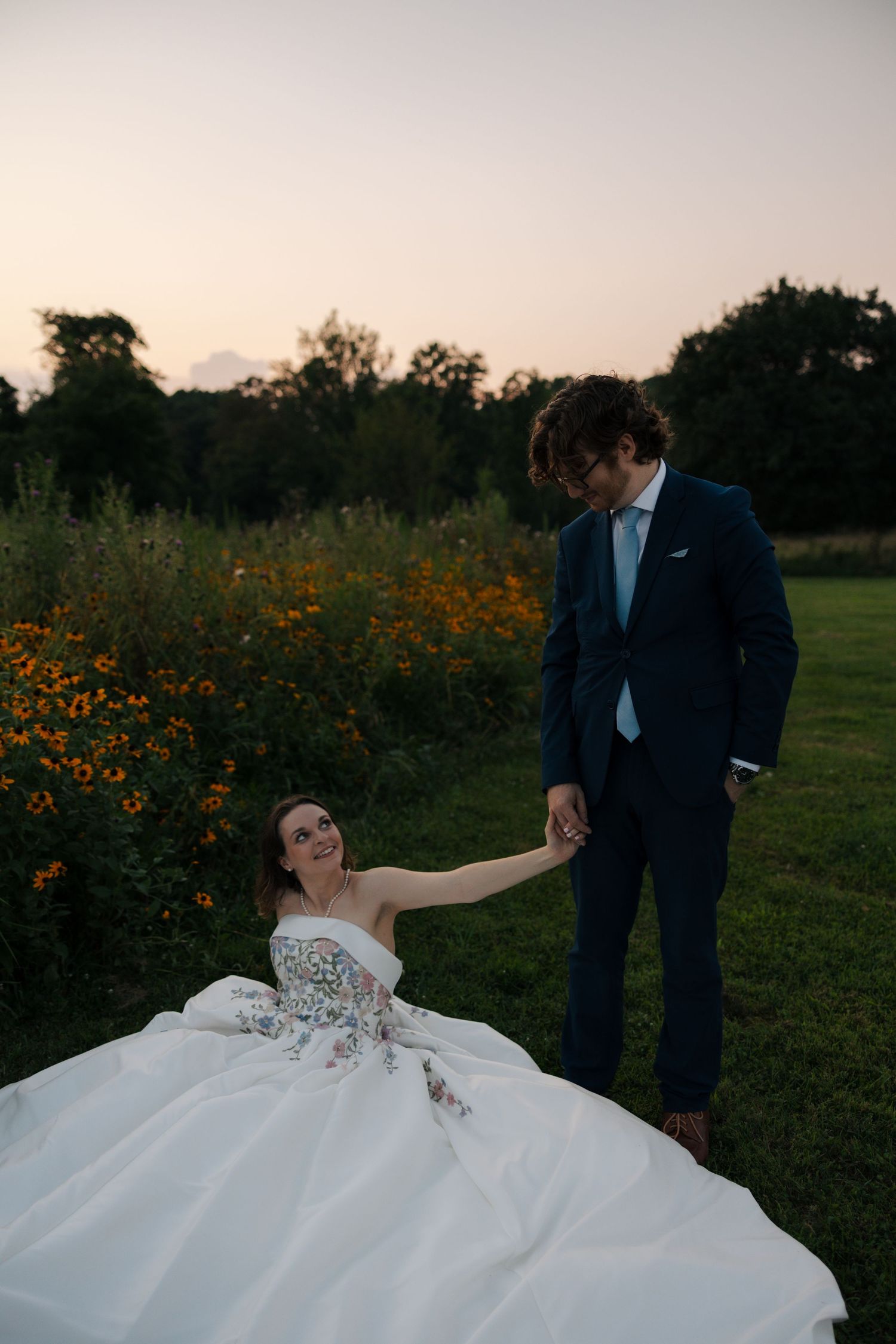Bride in strapless floral gown reaches up to hold grooms hand while seated in wildflower garden at dusk.