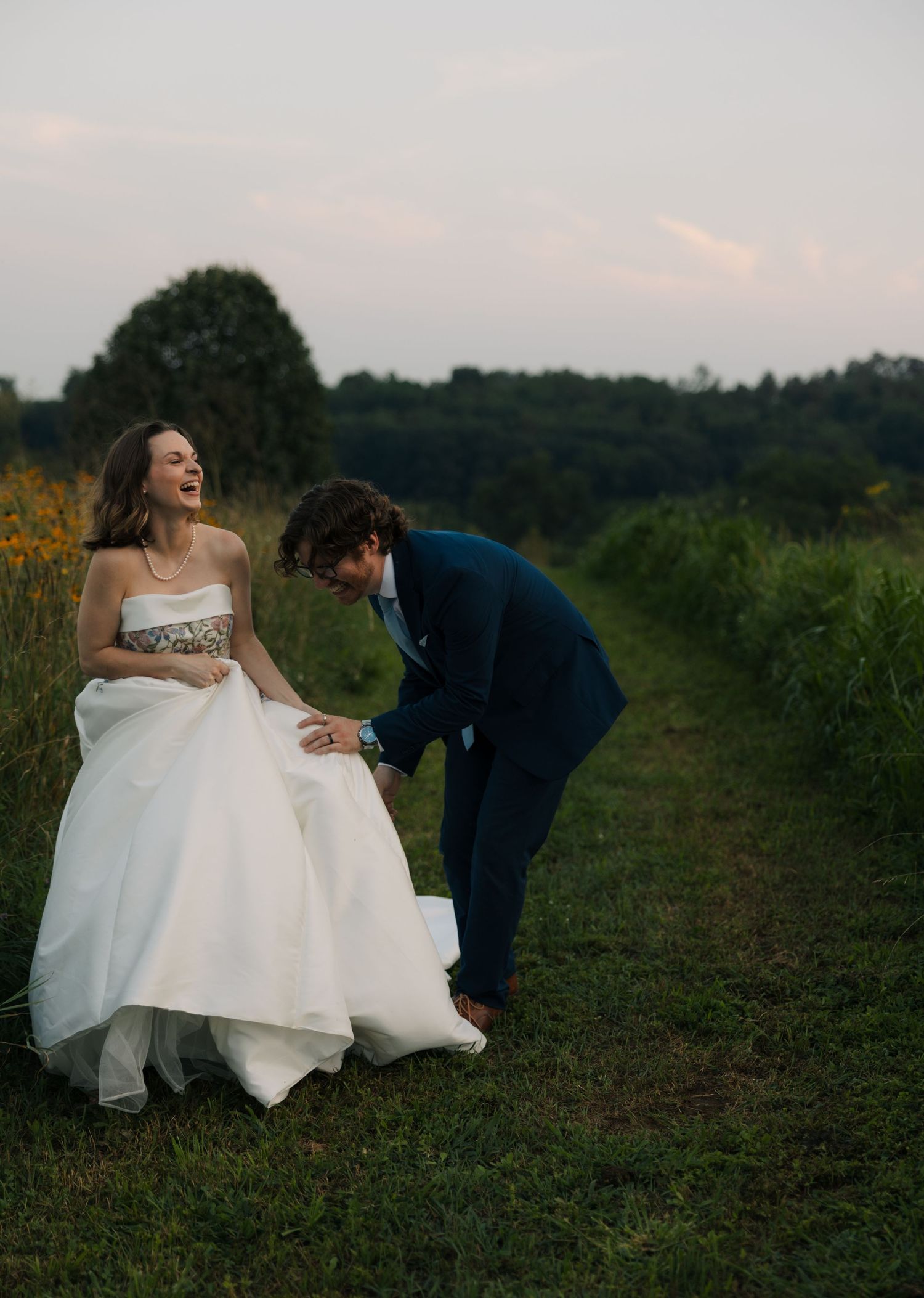 Laughing bride in white ballgown holds hands with groom in navy suit along a lush green meadow path.