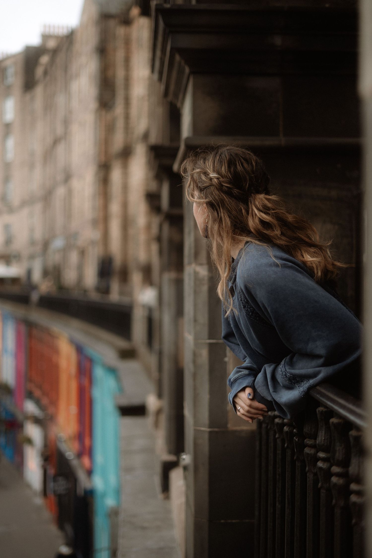 Woman in blue sweater leaning on iron railing of stone building balcony, gazing down colorful urban street.