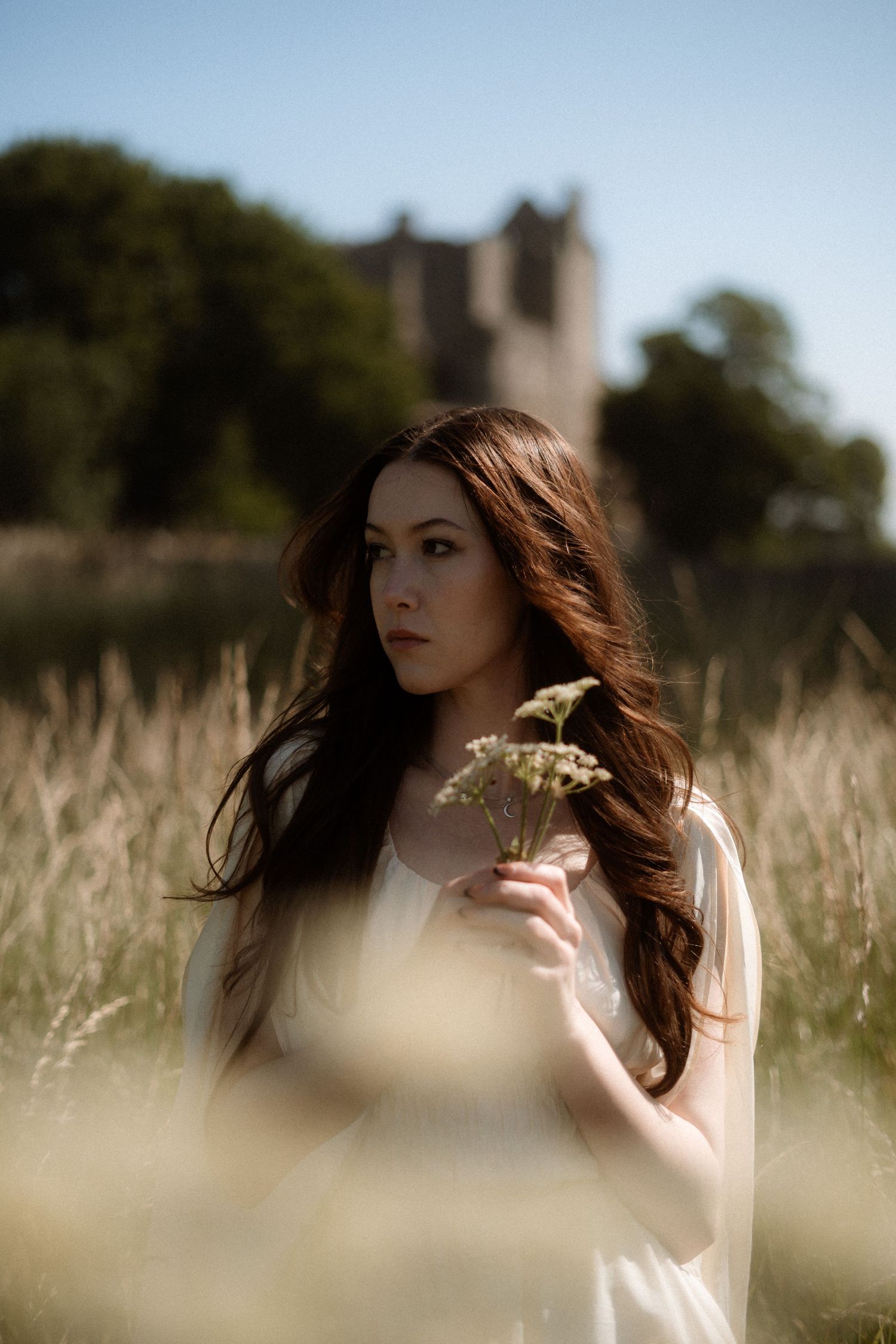 Ethereal photo of woman holding flowers, standing in tall grass with castle behind, Craigmillar Castle in Edinburgh, Scotland.