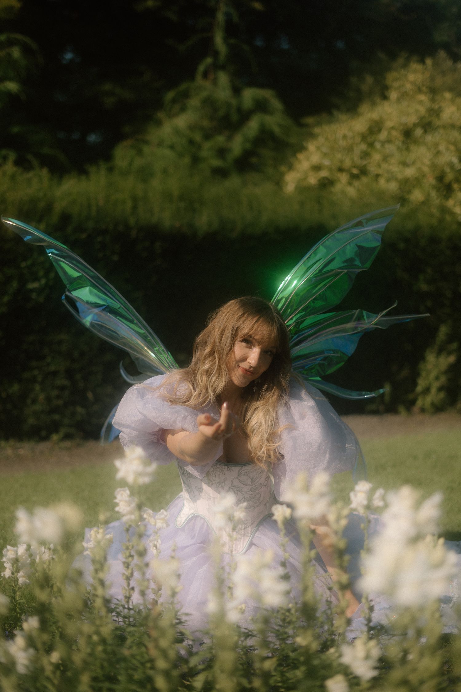 A figure in a white dress with iridescent fairy wings poses gracefully among blooming wildflowers in a dreamy garden setting.