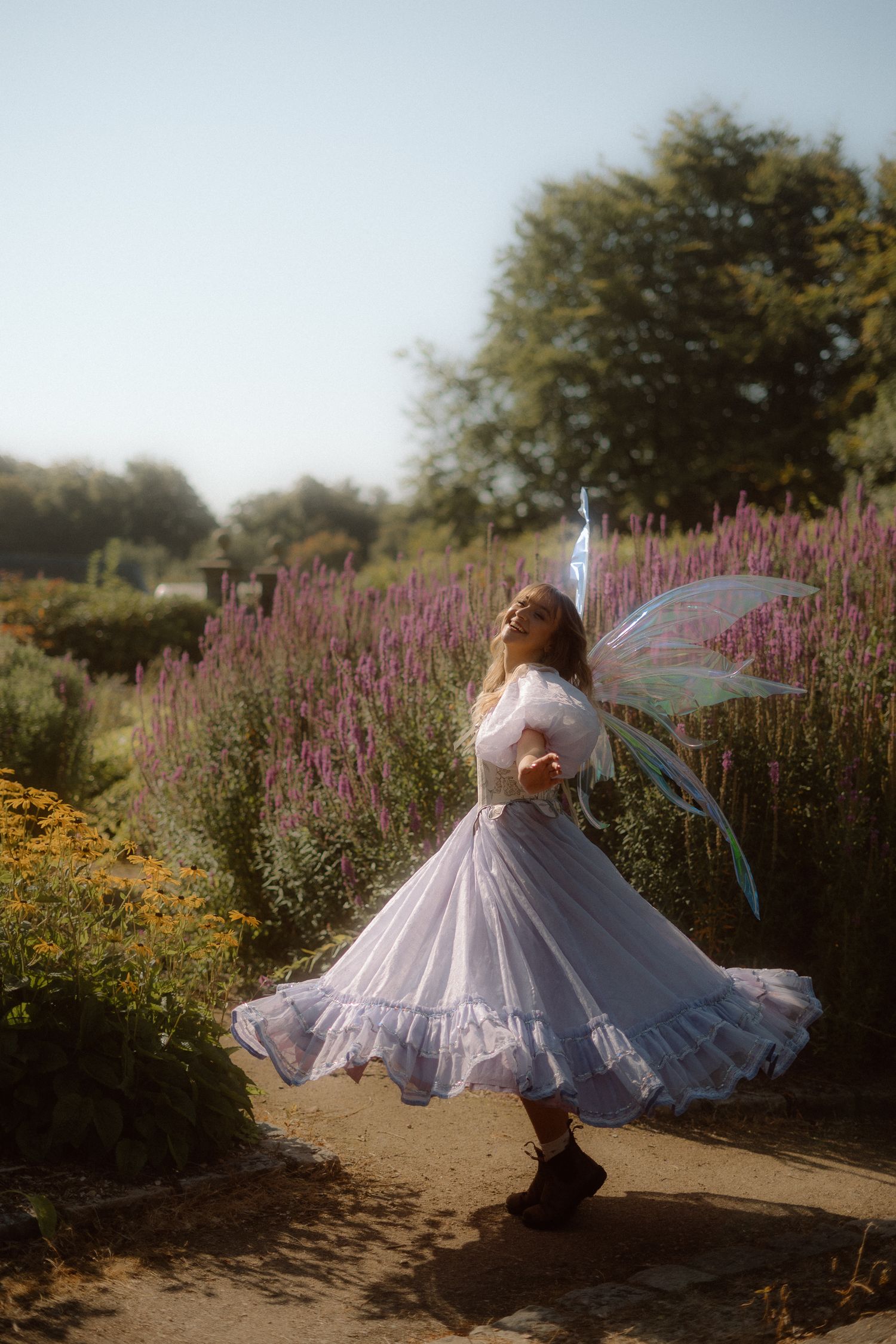 Person in flowing white dress twirling gracefully among purple wildflowers in a sunlit garden setting.