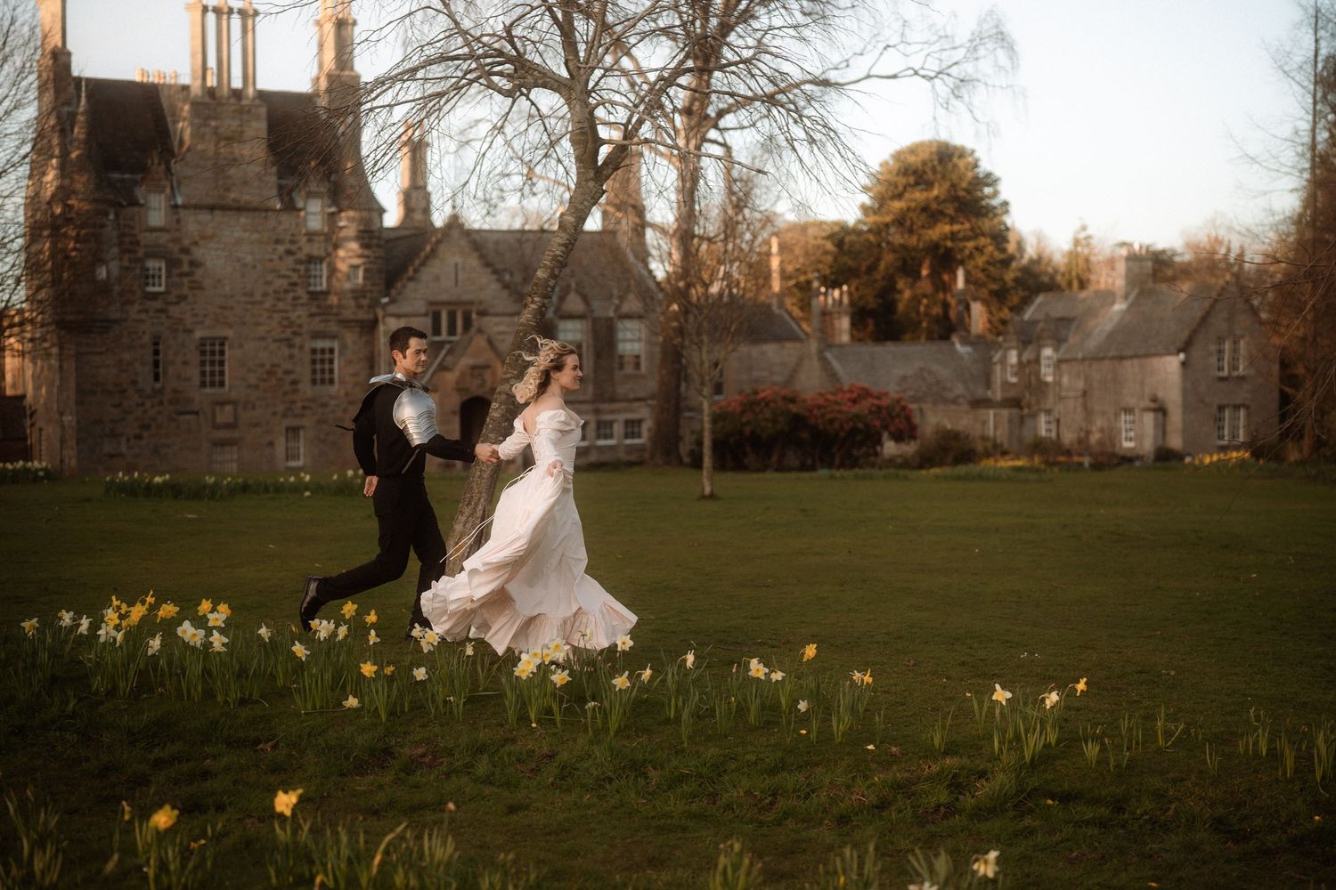 Bride and groom running through daffodil-lined lawn before a historic stone manor at golden hour.