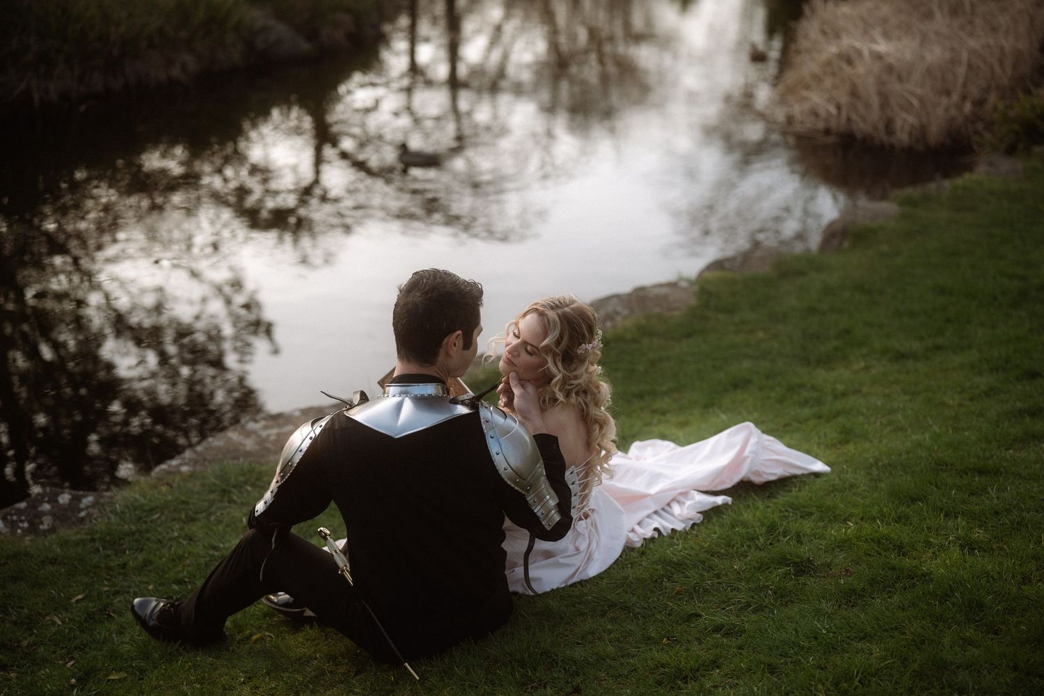 Bride and groom sitting together on green grass by a reflective pond, sharing a romantic moment on their wedding day.