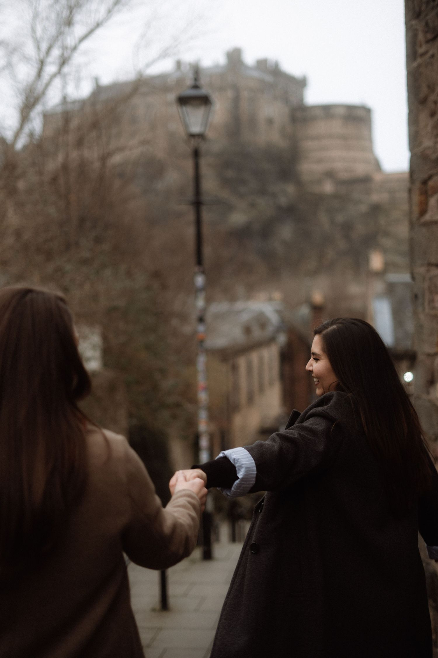 Two women holding hands walking toward Edinburgh Castle on a cobblestone street, one smiling back at the camera.