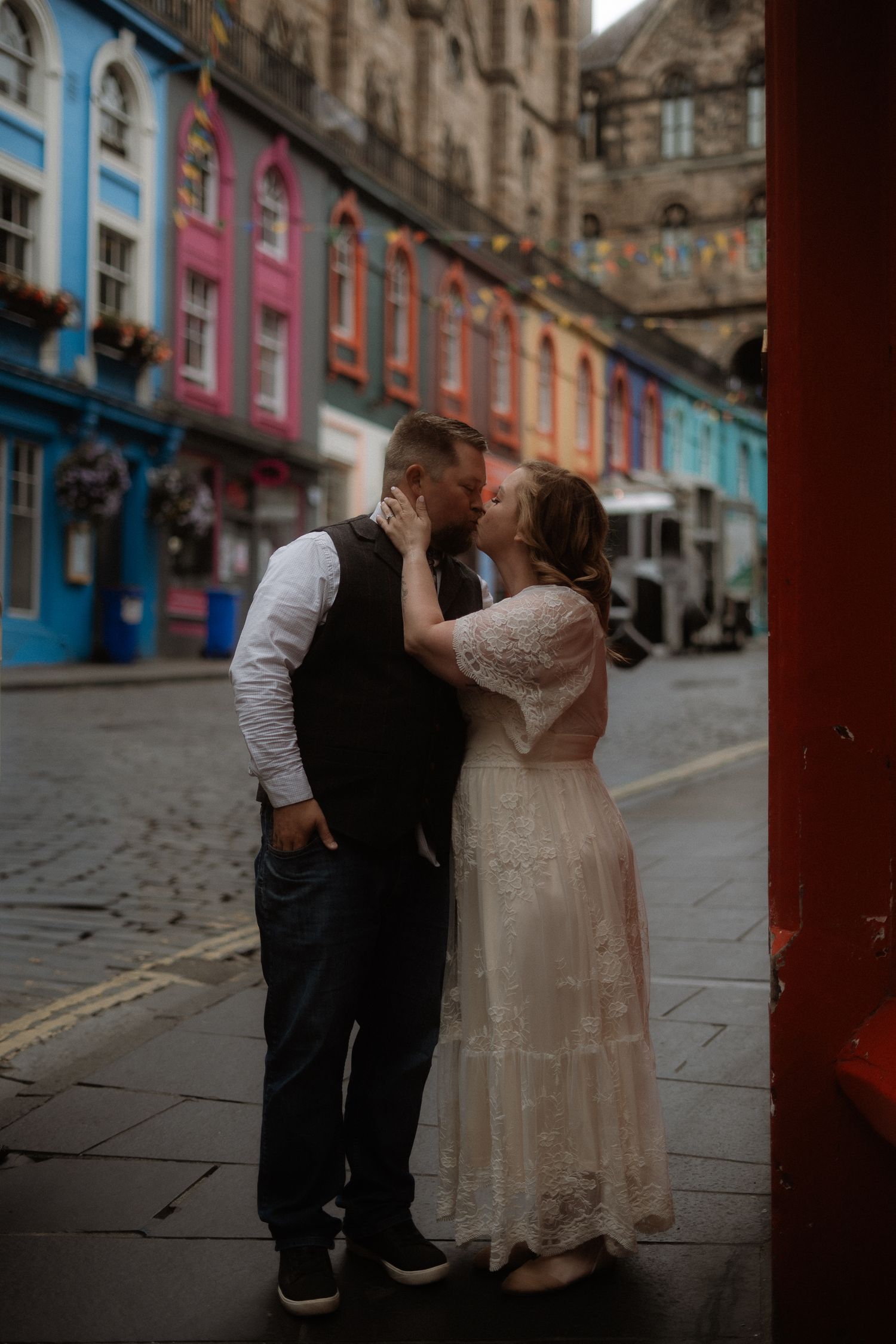 Couple sharing a romantic kiss on colorful Victoria Street in Edinburgh, Scotland, bride in lace dress.