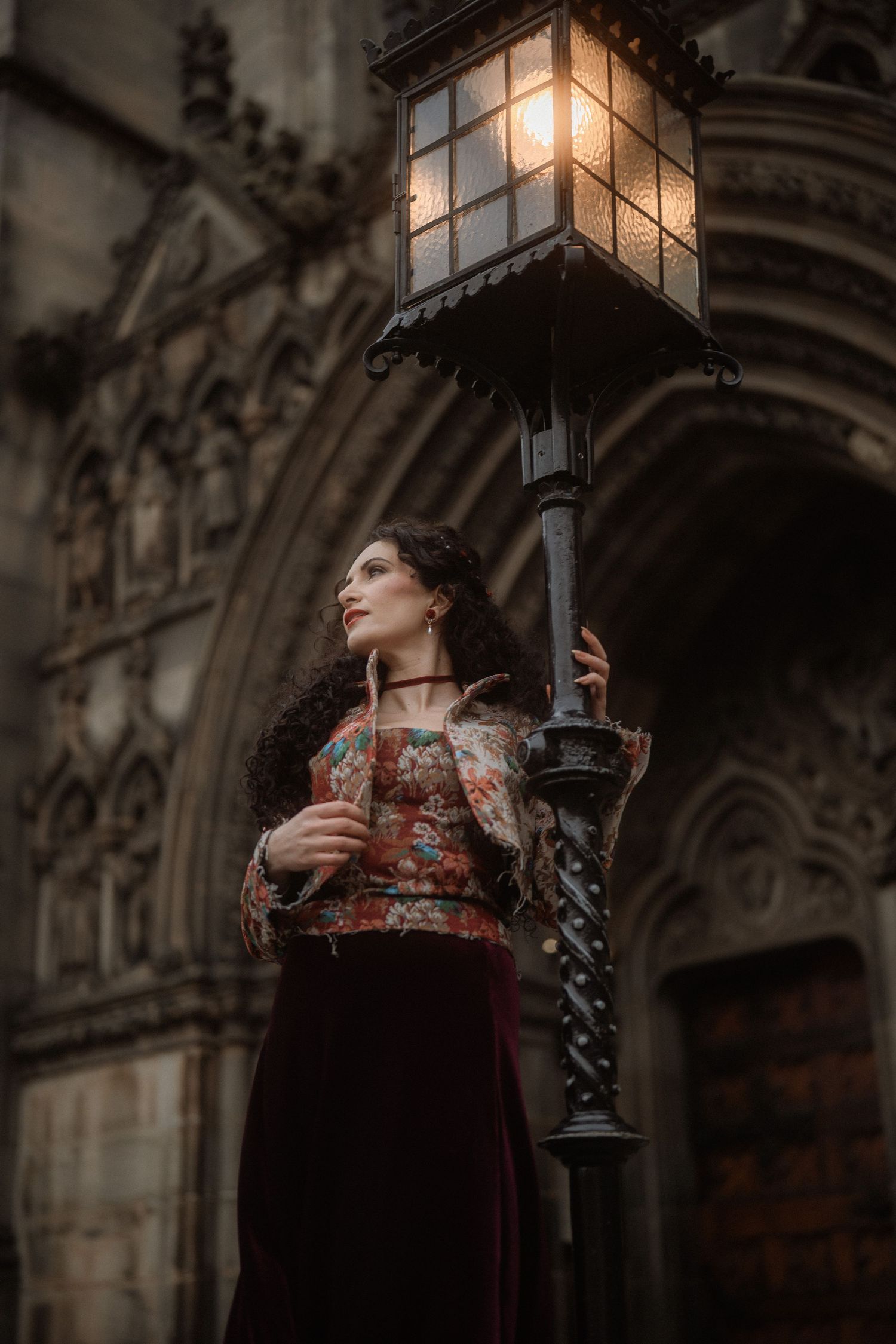 A woman in vintage floral dress holds a glowing street lamp outside a Gothic stone cathedral at dusk.