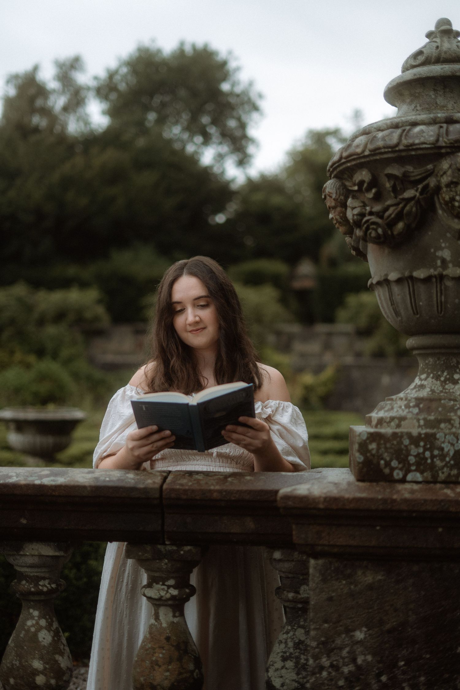 Women dressed in white dress leaning on stone arch in an Italian garden, reads a book. Bridgeton and pride and prejudice vibes. Pollock Park