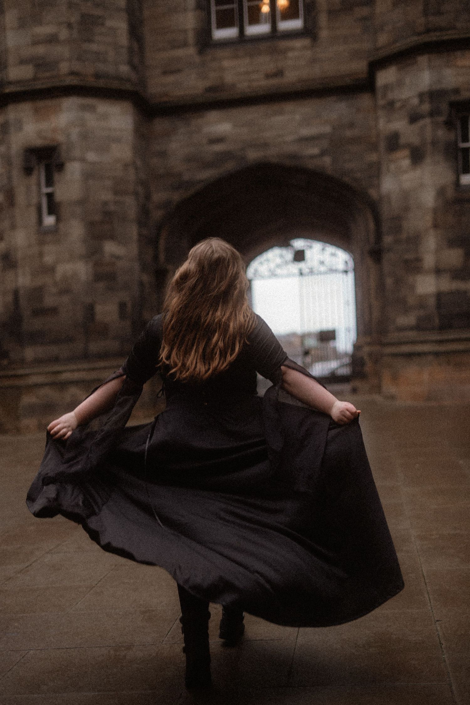 A person in a flowing black dress twirls beneath a stone archway of a historic building.