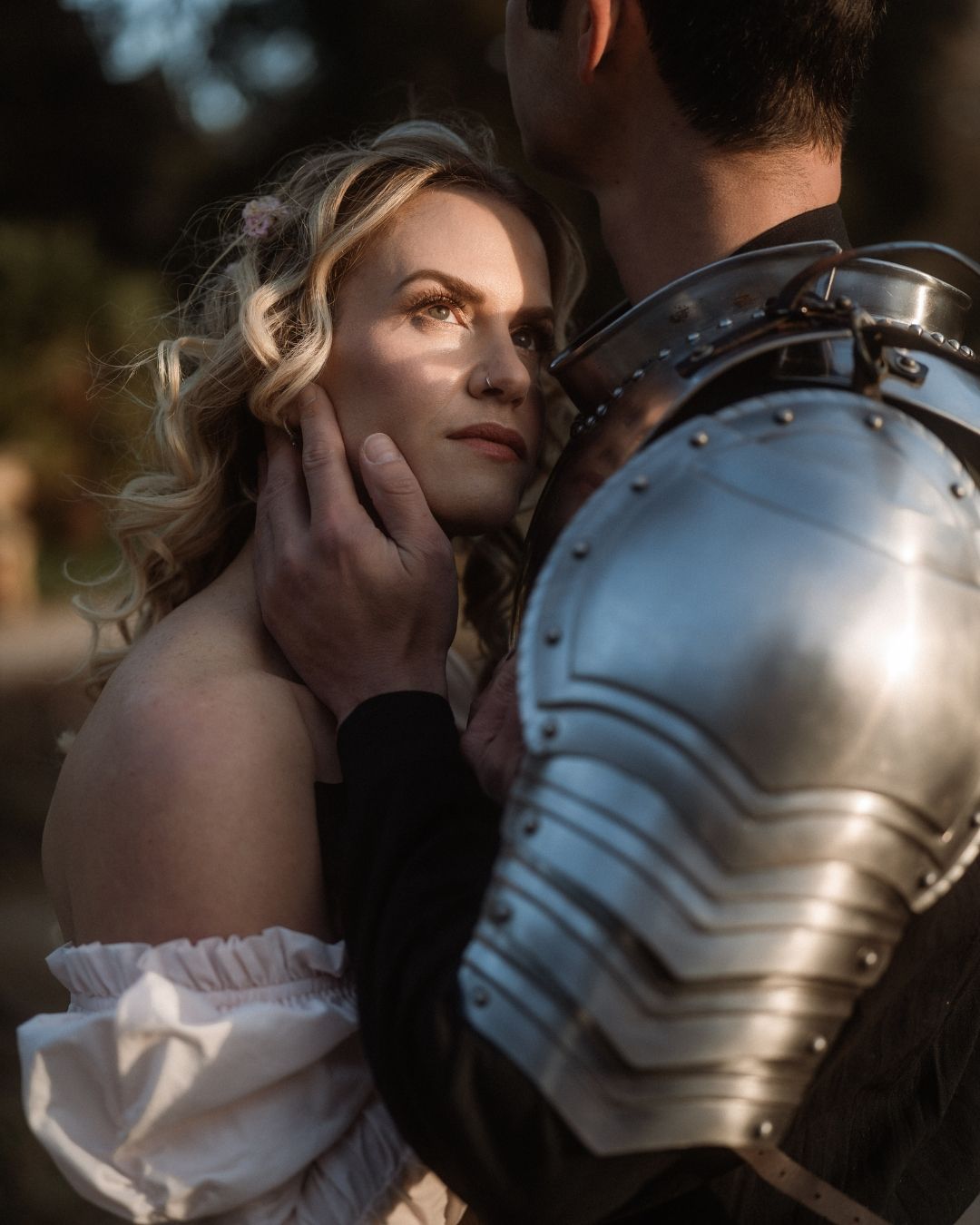 A knight in armor tenderly cups a blonde woman's face in a romantic medieval-themed portrait shoot.