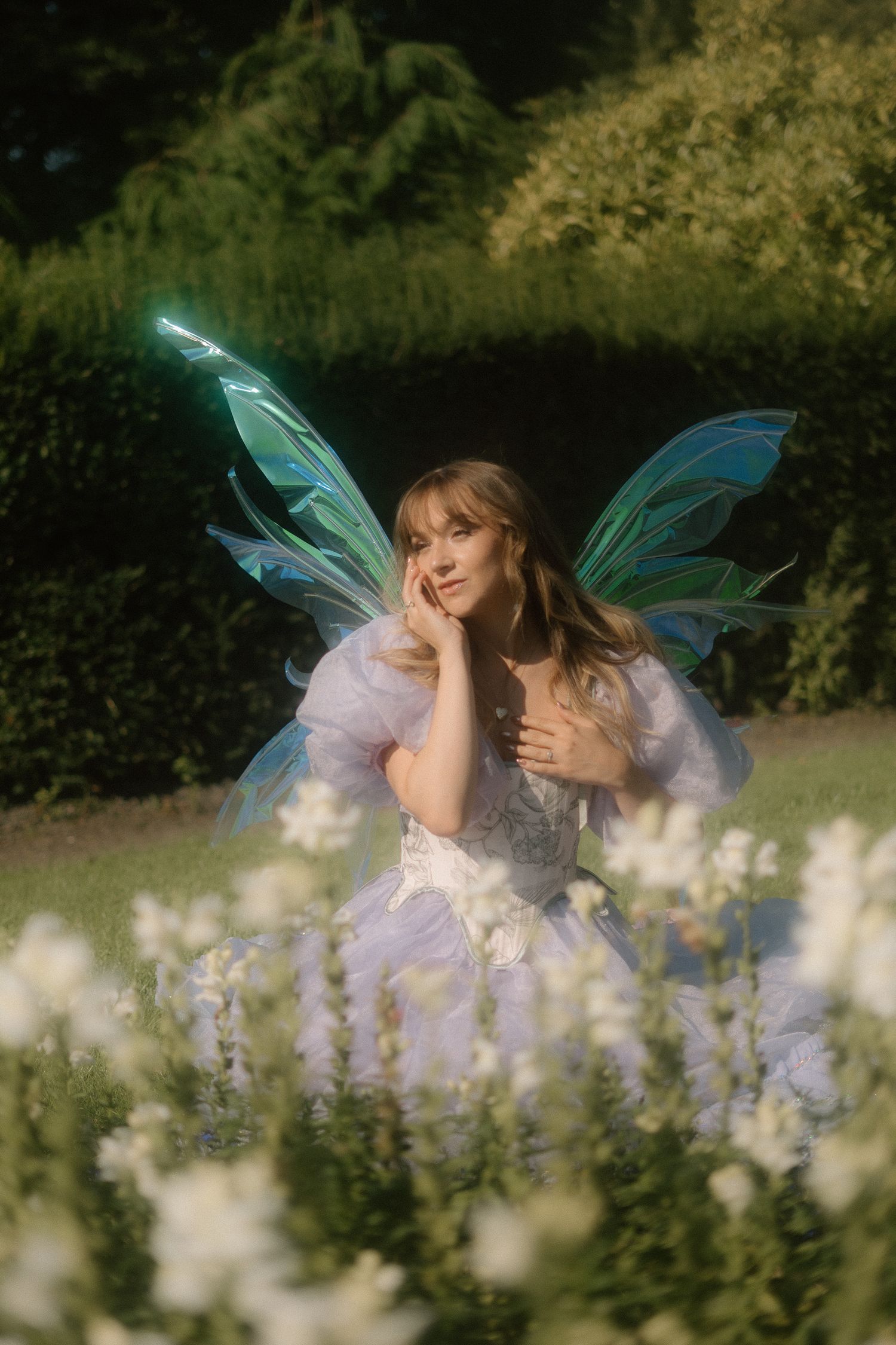 A fairy with iridescent wings poses dreamily in a garden of white flowers wearing a flowing lilac dress.