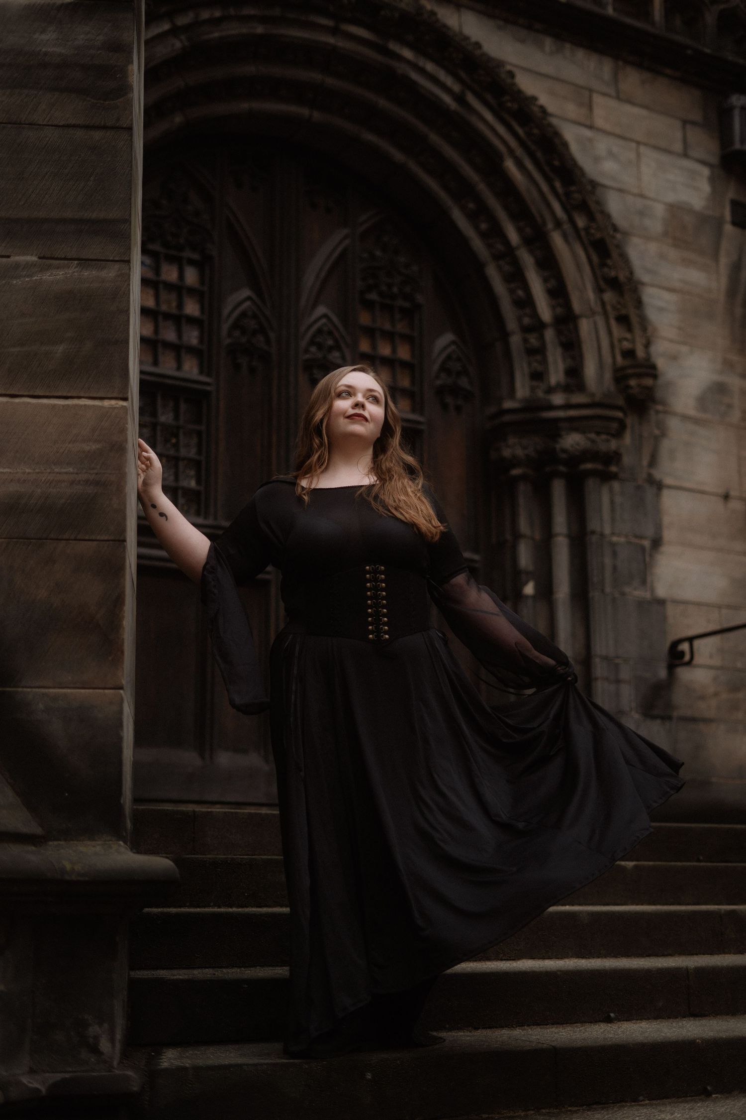A figure in a flowing black gothic dress poses dramatically on stone steps outside an arched medieval church doorway.