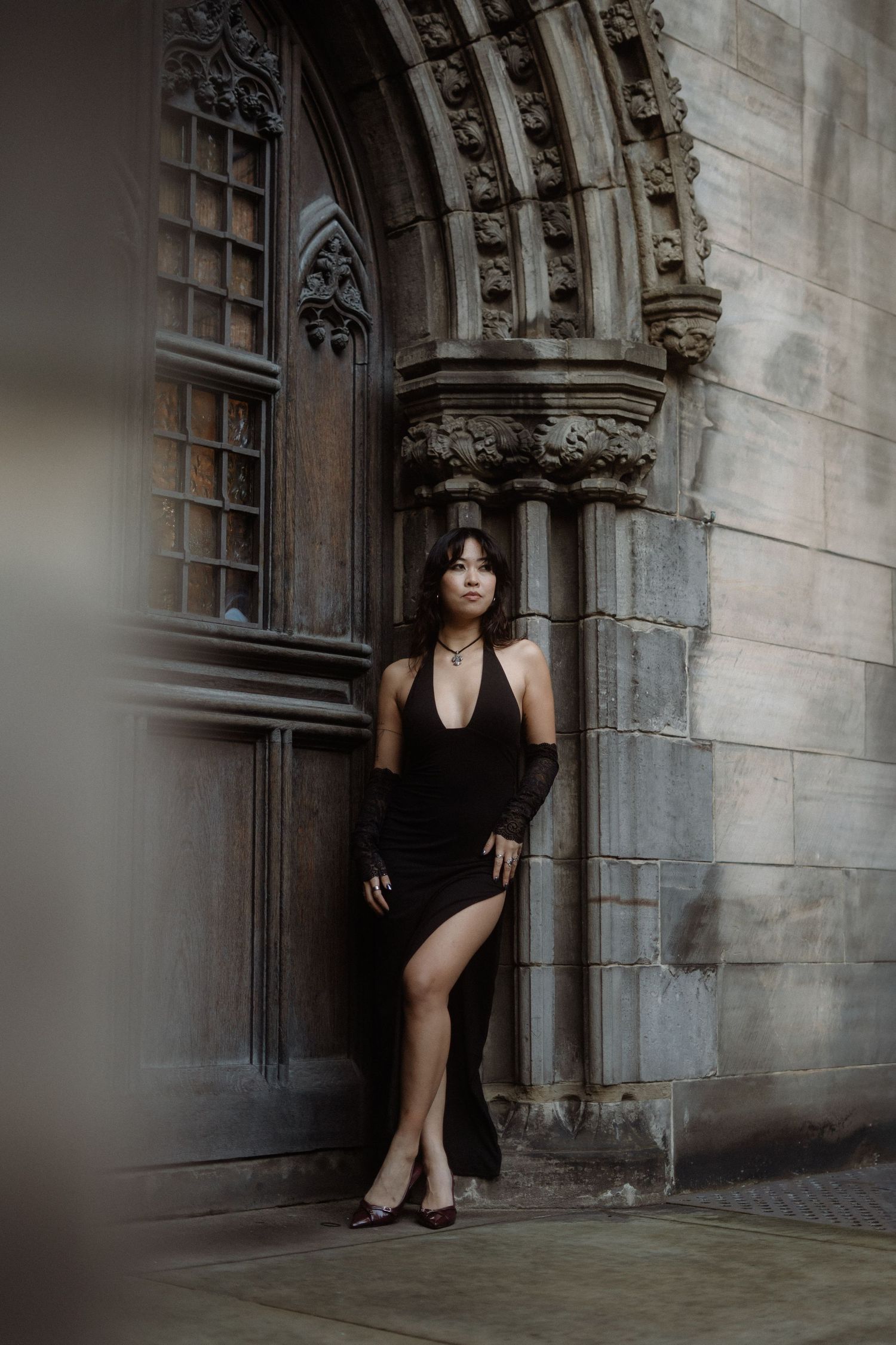 A person in a black dress poses dramatically in a Gothic stone doorway of an old church or cathedral.