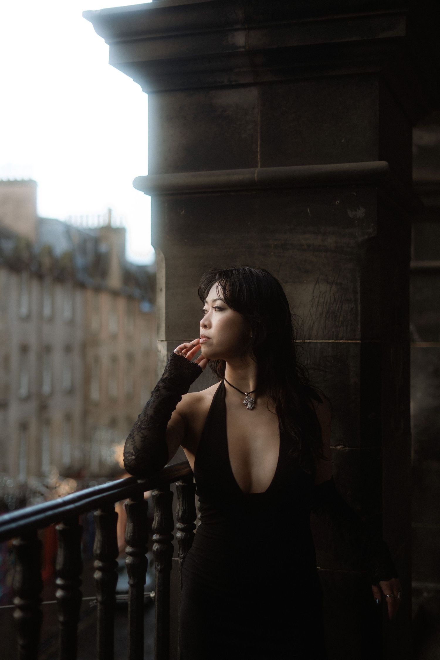 A woman in a black dress stands thoughtfully on a balcony with historic architecture in the background.
