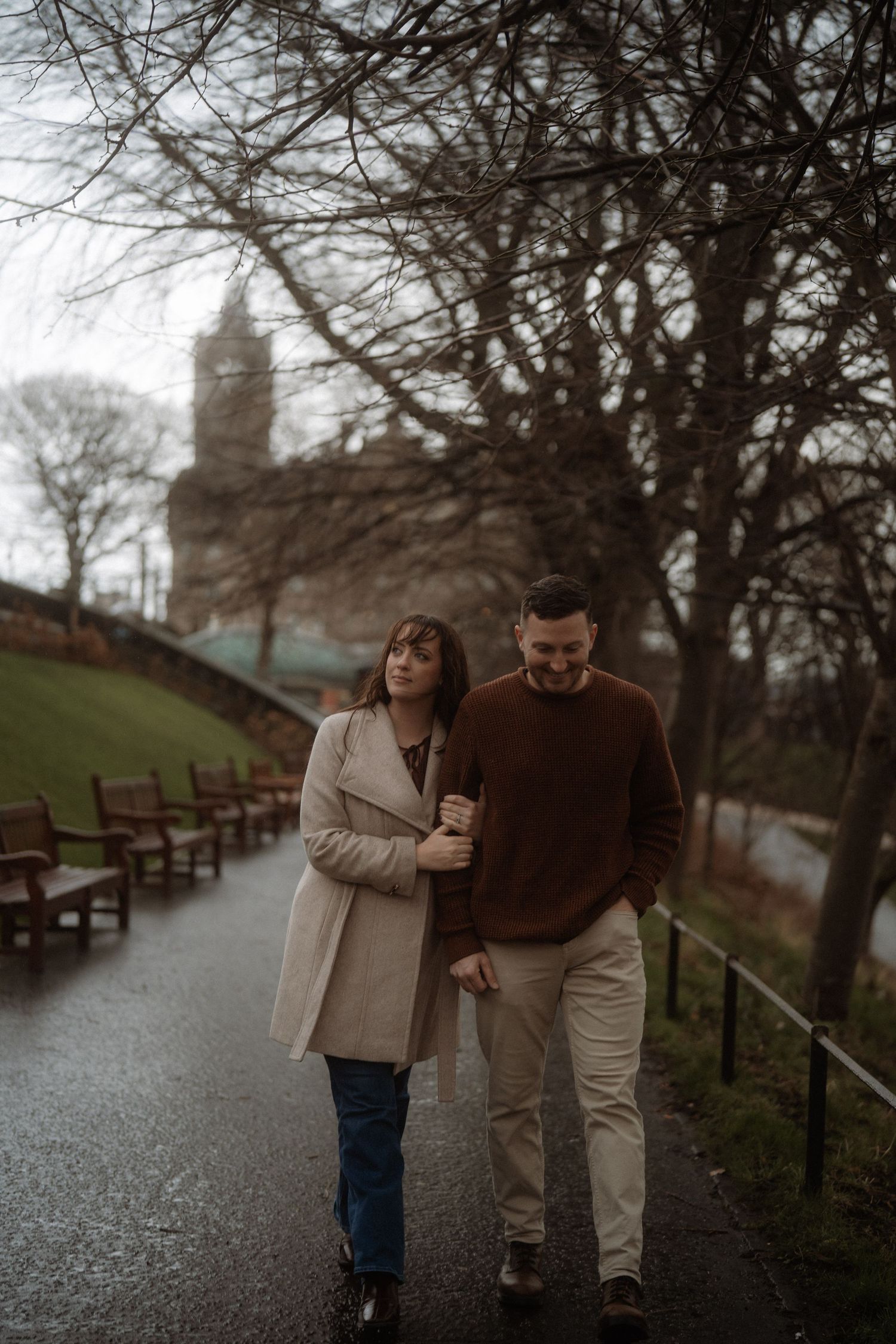 Couple walking arm in arm on a rainy park path, woman in beige coat and man in brown sweater, historic tower behind.