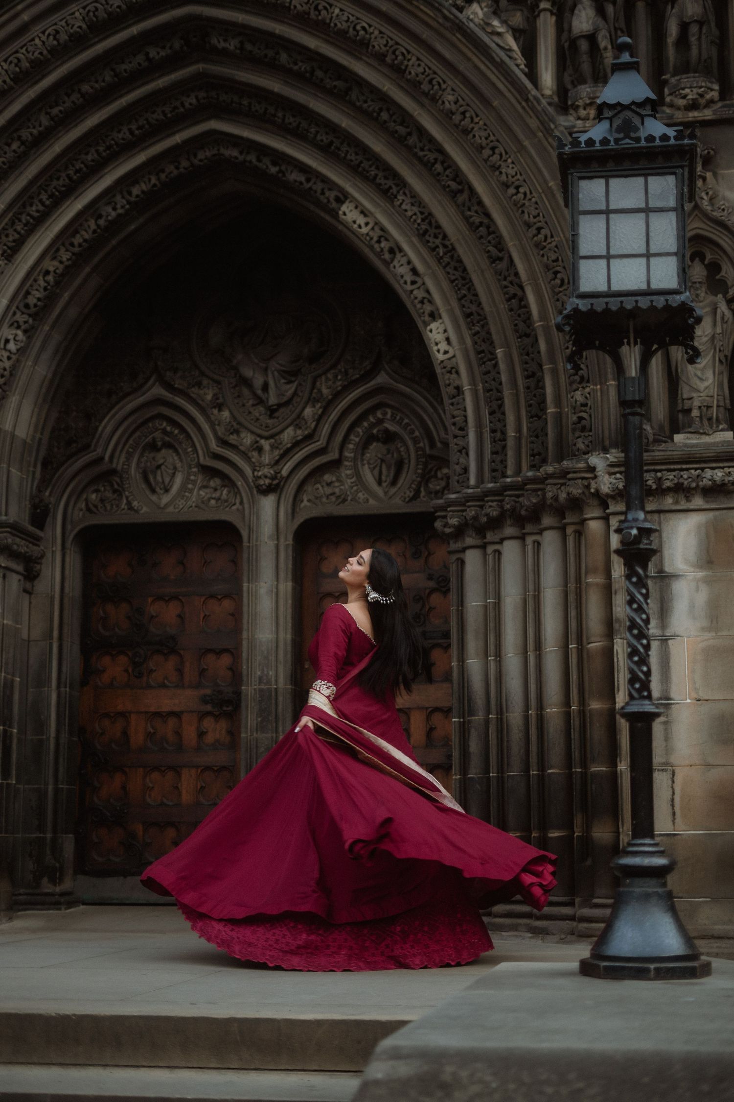 A flowing burgundy dress twirls dramatically in front of ornate Gothic cathedral doors.