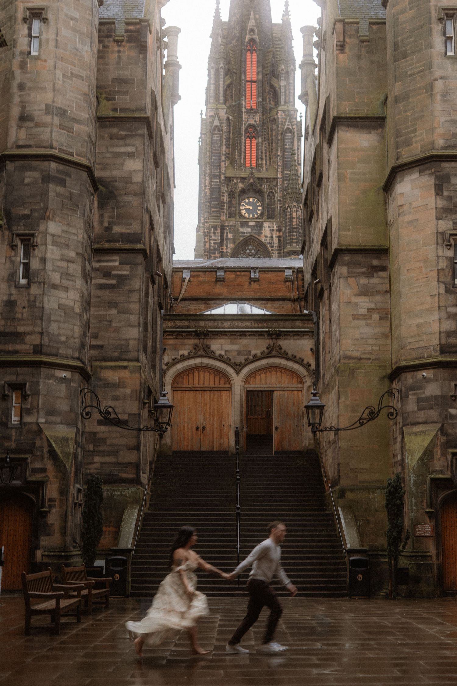 Couple holding hands running through a grand stone cathedral courtyard with Gothic architecture and clock tower backdrop.